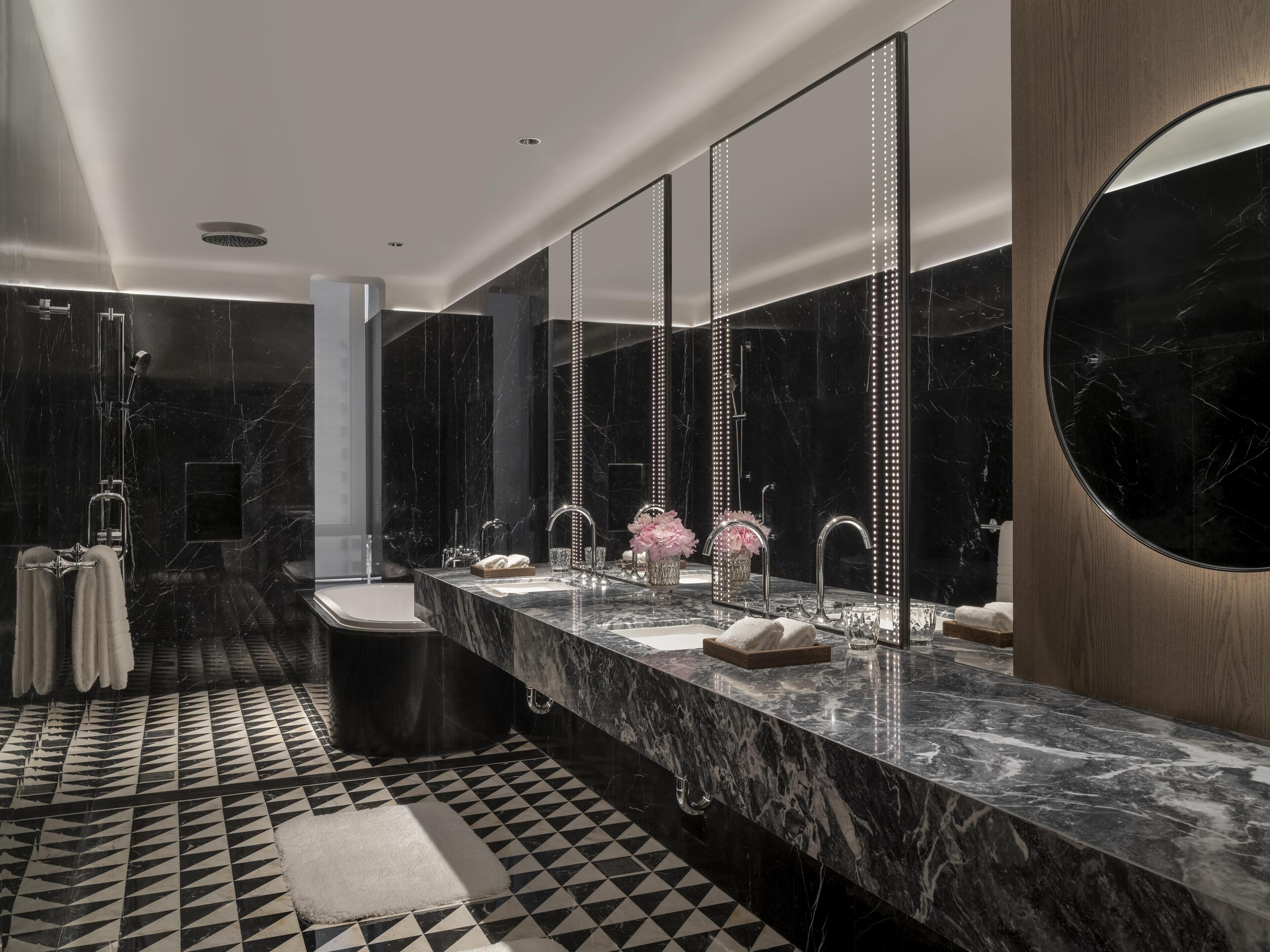 Un baño moderno con piso a cuadros blancos y negros, un tocador de mármol con tres lavamanos y un espejo grande con luces LED verticales.