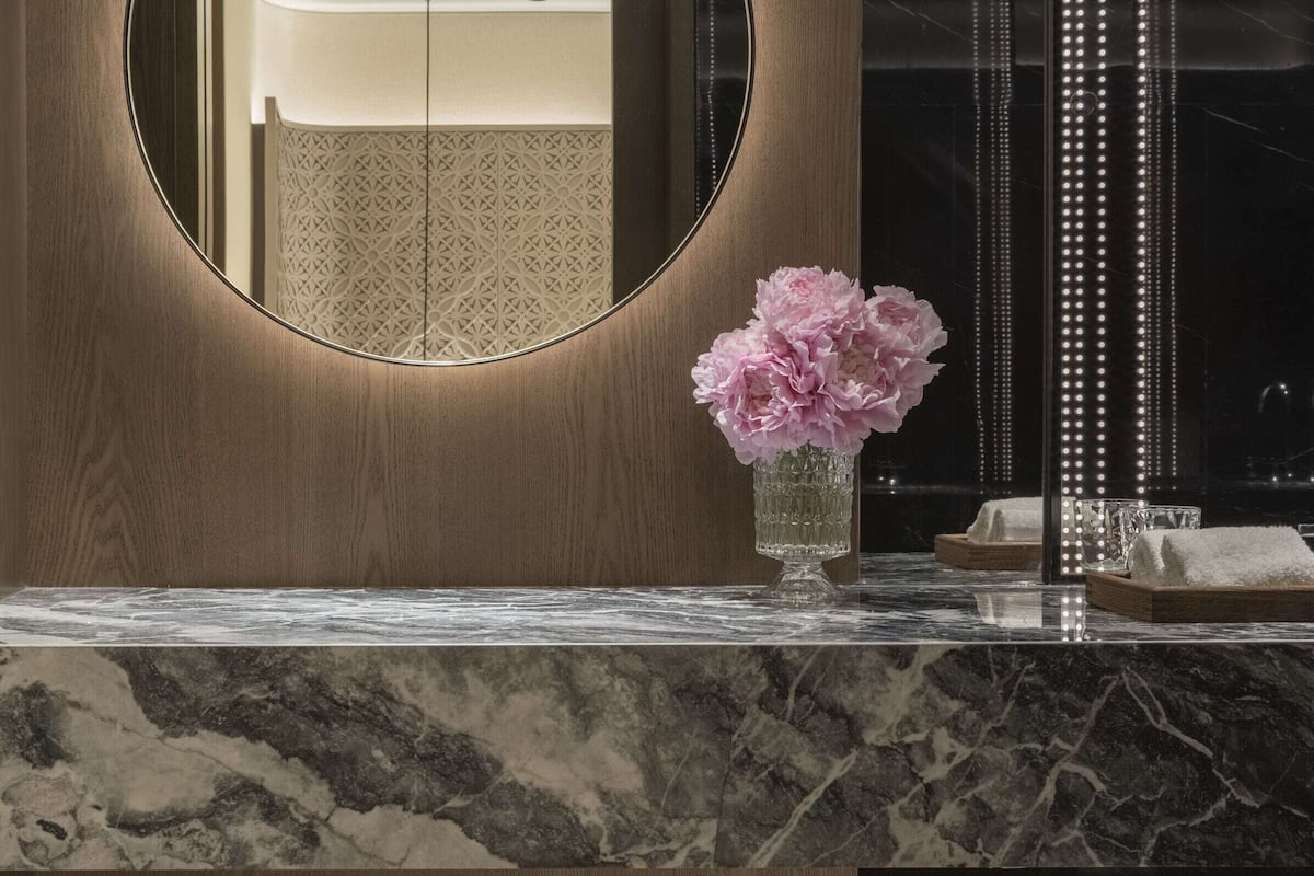A bathroom with a wooden vanity, a round mirror, a vase with flowers, and a vertical LED light strip.
