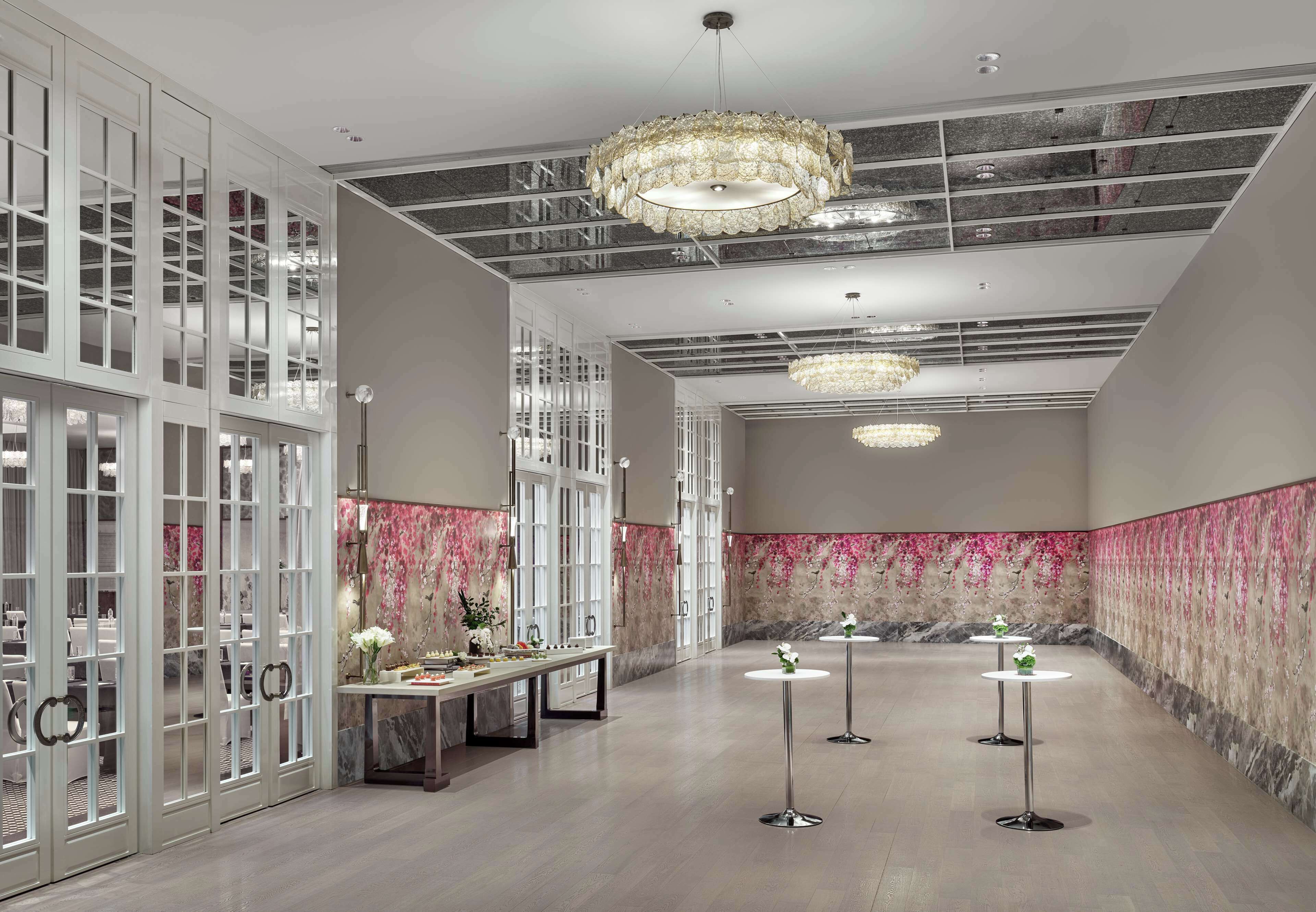 A spacious hotel lobby with high ceilings, large chandeliers, and a marble wall with pink floral wallpaper.