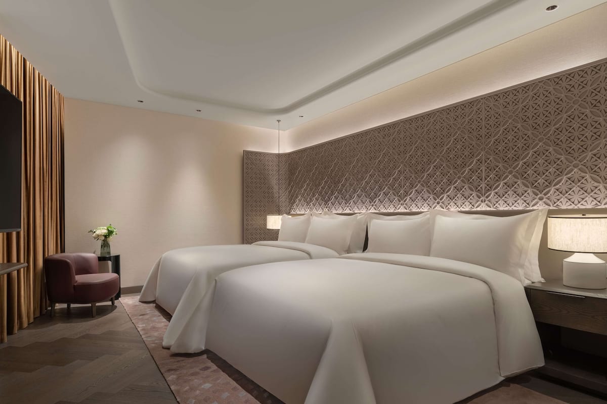 A modern hotel room with two beds, a sitting area with a chair, and a decorative wall panel.