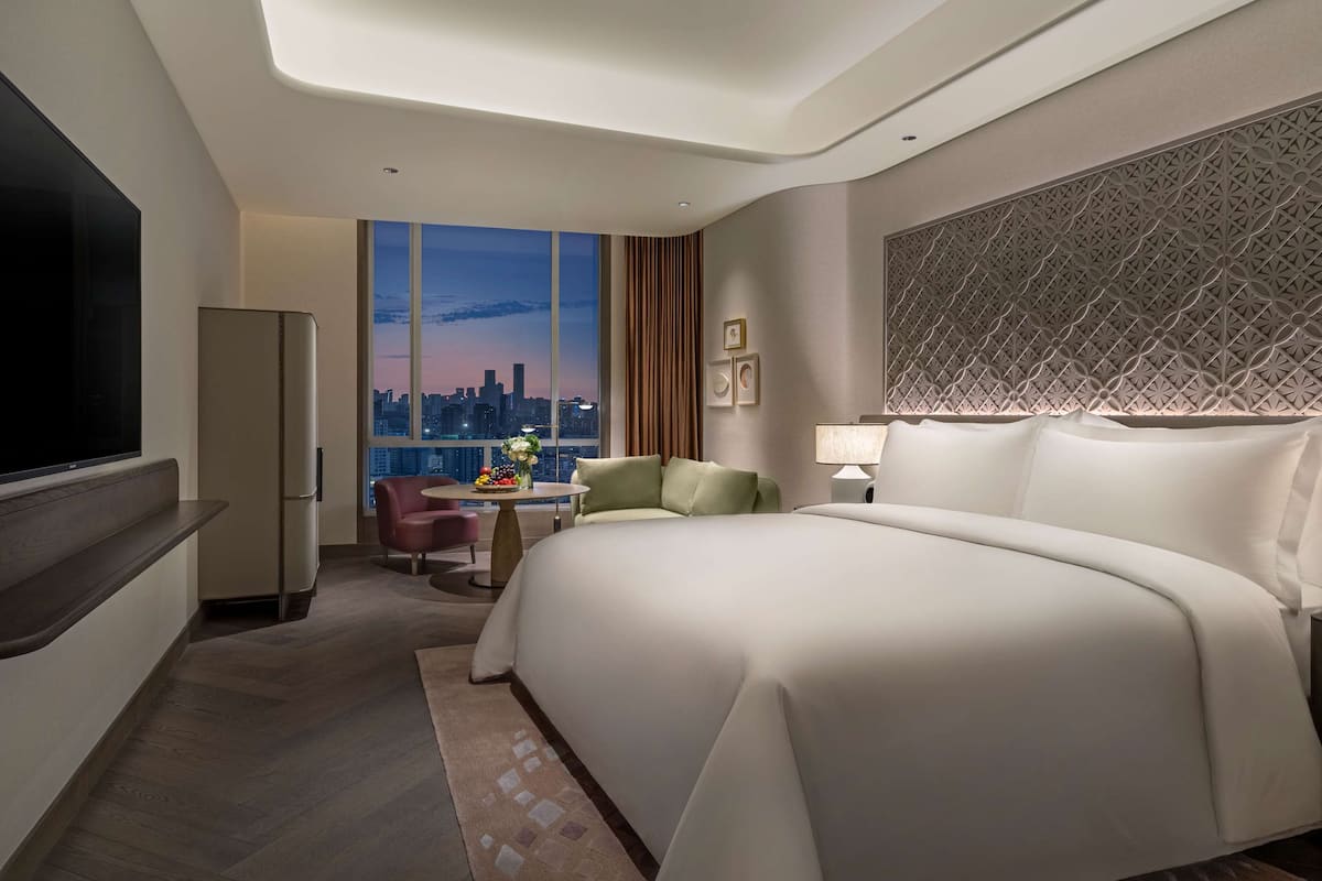 A modern hotel room with a large bed, a flat-screen TV, a dining table with a vase of flowers, and a city view through large windows.