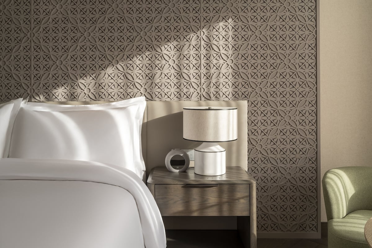 A hotel room with a bed, a nightstand with a lamp, a clock, and a green armchair.