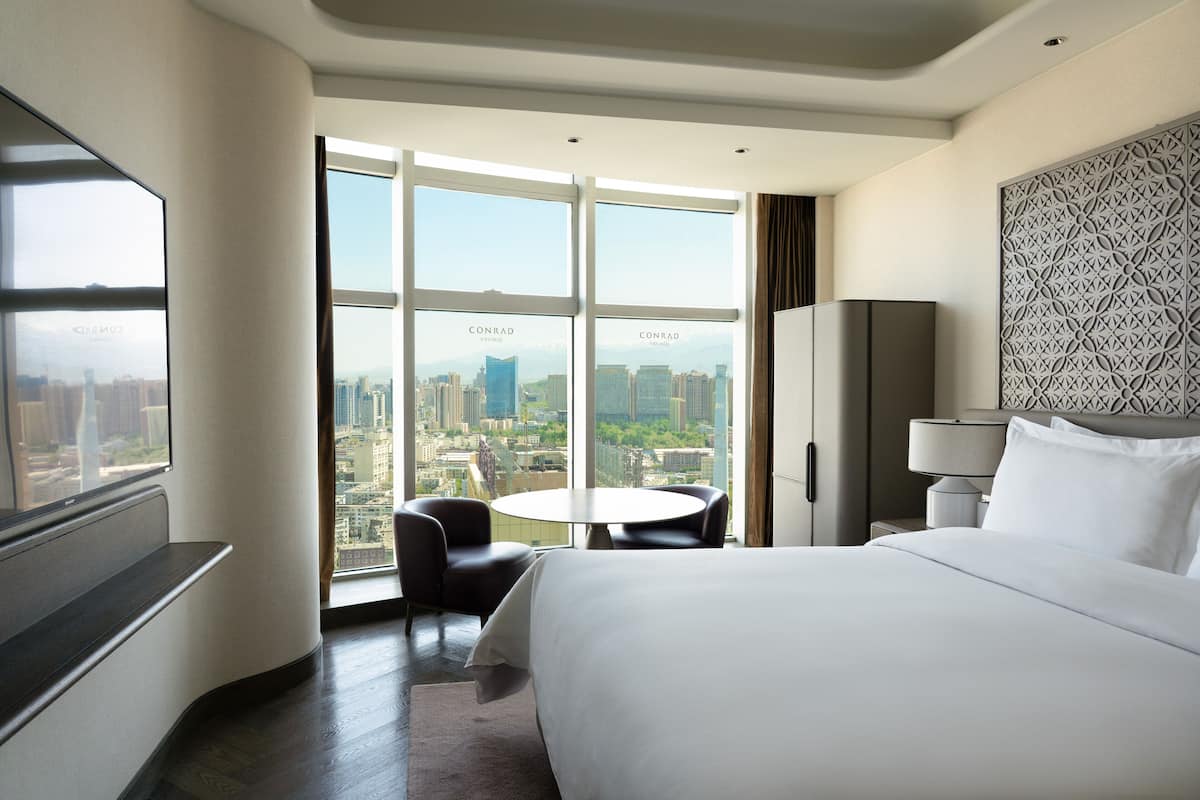 A hotel room with a large bed, a round table, two chairs, a TV, and a view of a cityscape through large windows.
