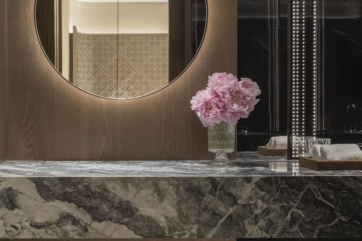 A bathroom with a wooden vanity, a round mirror, a vase with flowers, and a vertical light strip.