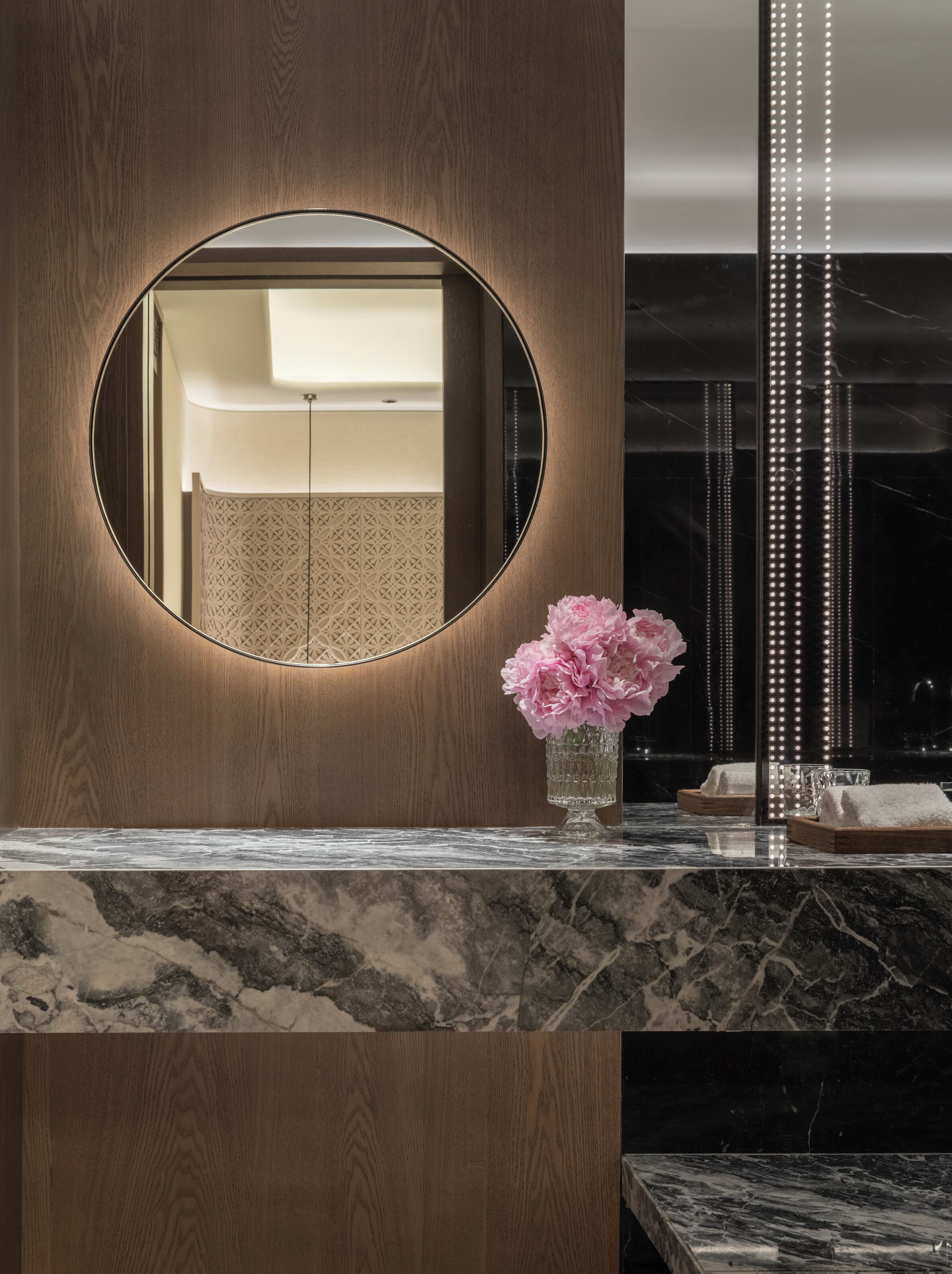 Ein Badezimmer mit einem Holzkosmetikschrank, einem runden Spiegel und einer Vase mit rosa Blumen.