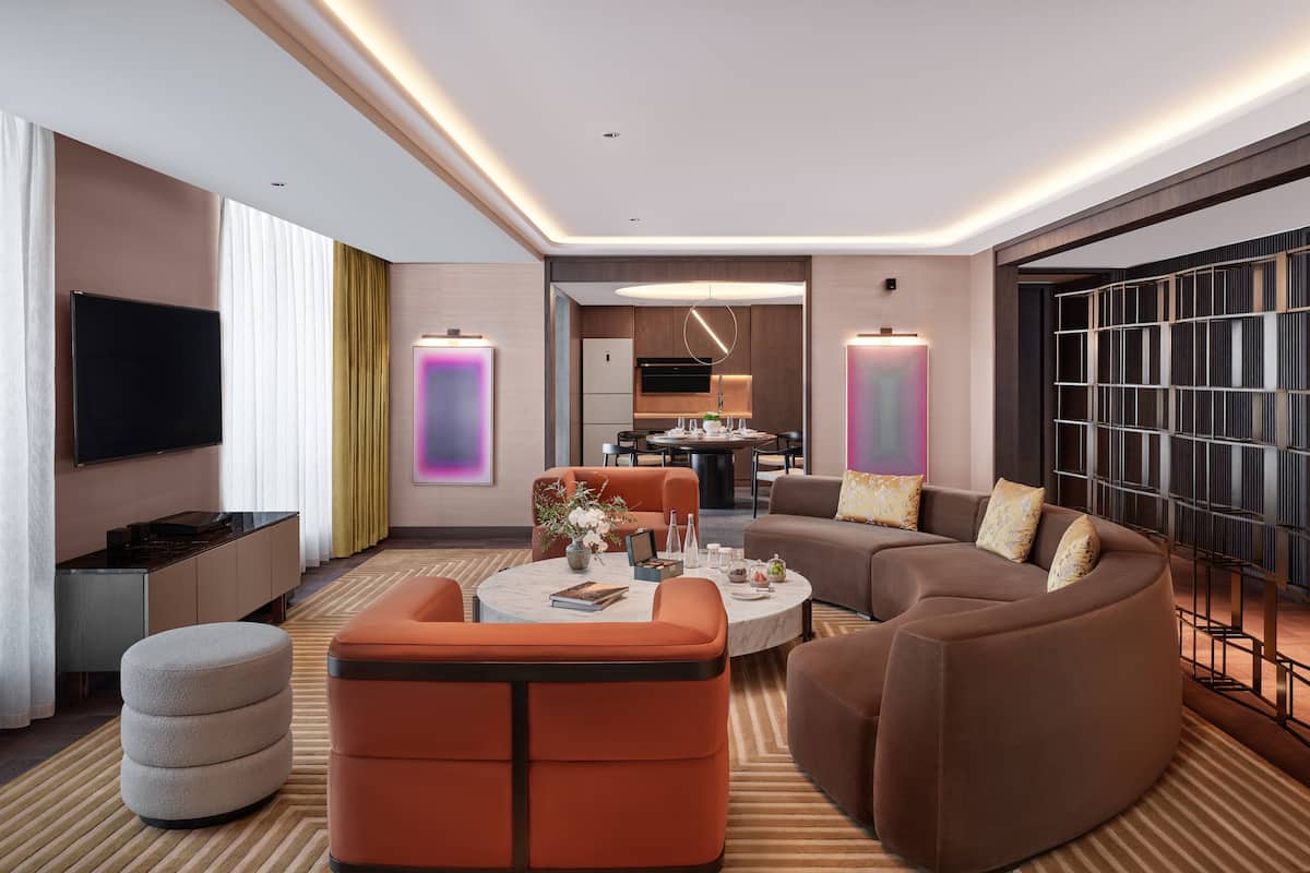 A modern hotel room with a living area featuring a sofa, ottoman, and coffee table. There is a flat-screen TV mounted on the wall, a dining table with chairs, and a kitchen area visible through an open door.