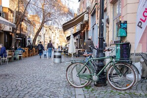 A cobblestone street lined with cafes and outdoor seating, a green bicycle parked by the curb, and a trash can in the foreground. - Dafi (Plovdiv)