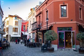 A cobblestone street with buildings, outdoor seating, and a storefront with a visible sign. - Dafi (Plovdiv)