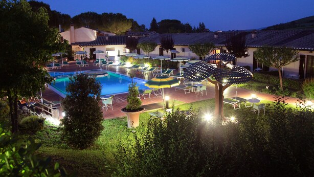 Zona de piscina con sillas de descanso y sombrillas, rodeada de vegetación y edificios.