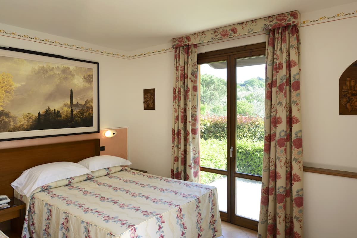 A hotel room with two beds, a large landscape painting, a wooden headboard, a window with floral curtains, and a small wooden artwork on the wall.