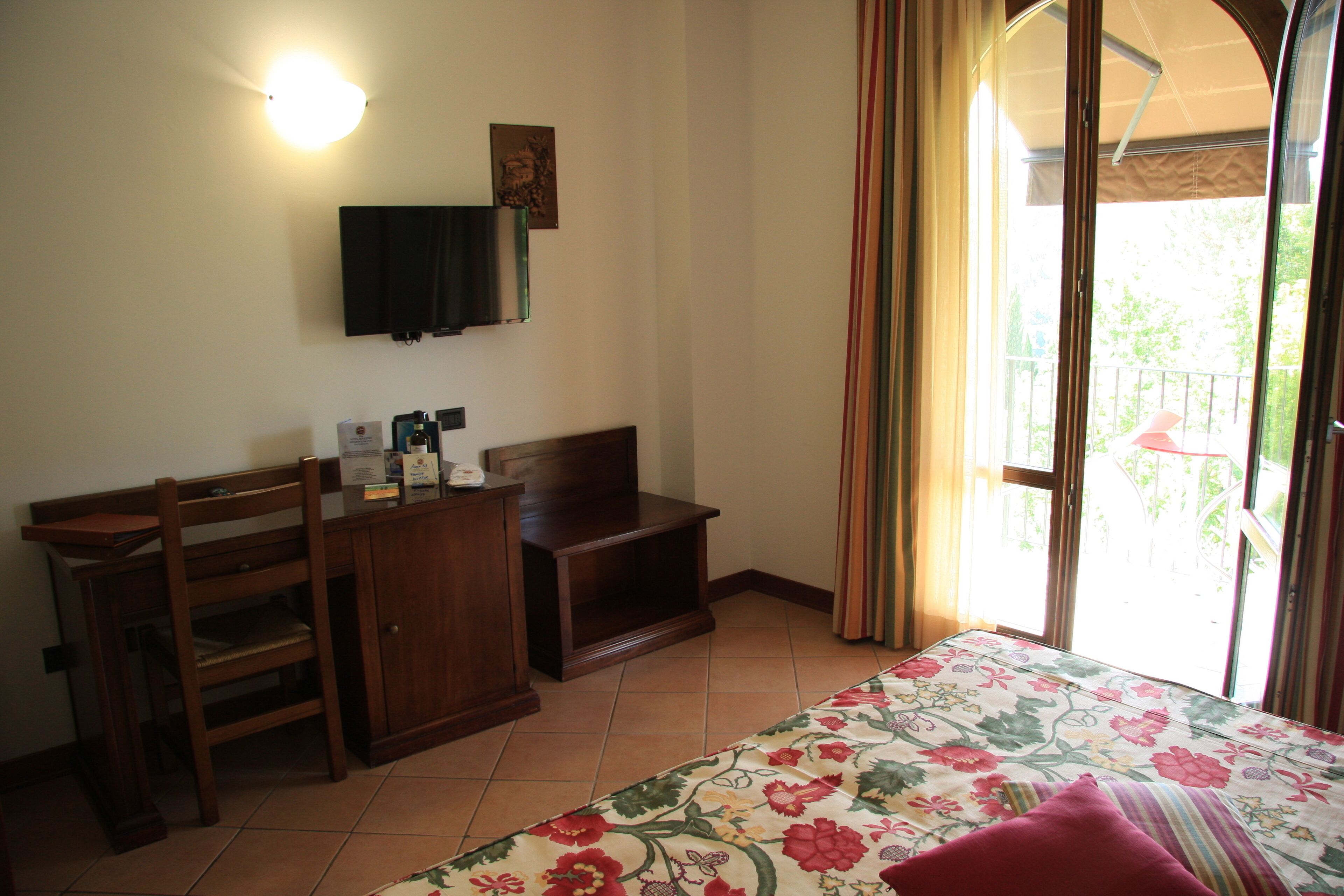 Un dormitorio con cama, escritorio, silla, televisor y balcón con cortinas.
