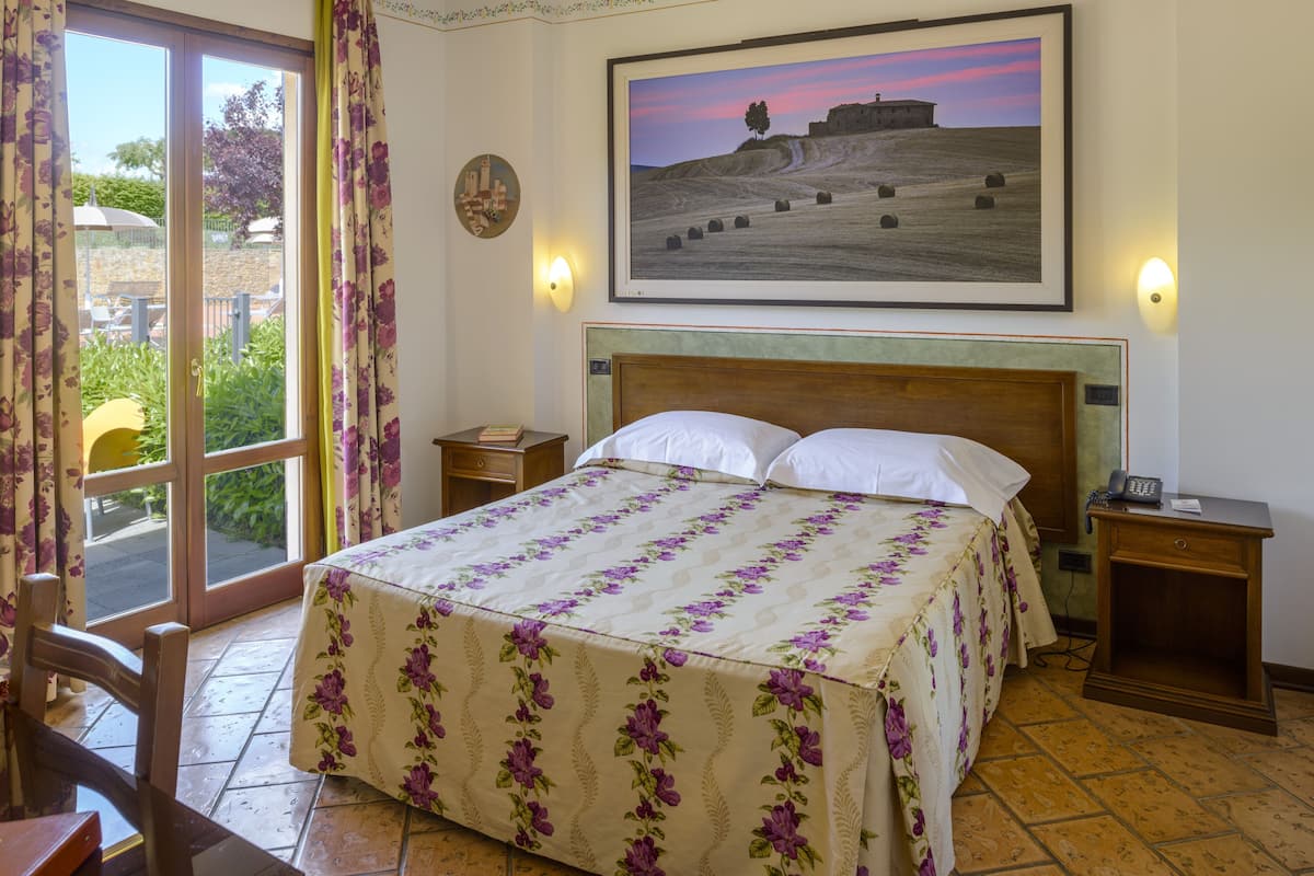A hotel room with a bed, a nightstand, a chair, a table, a painting on the wall, and a view of the outside.