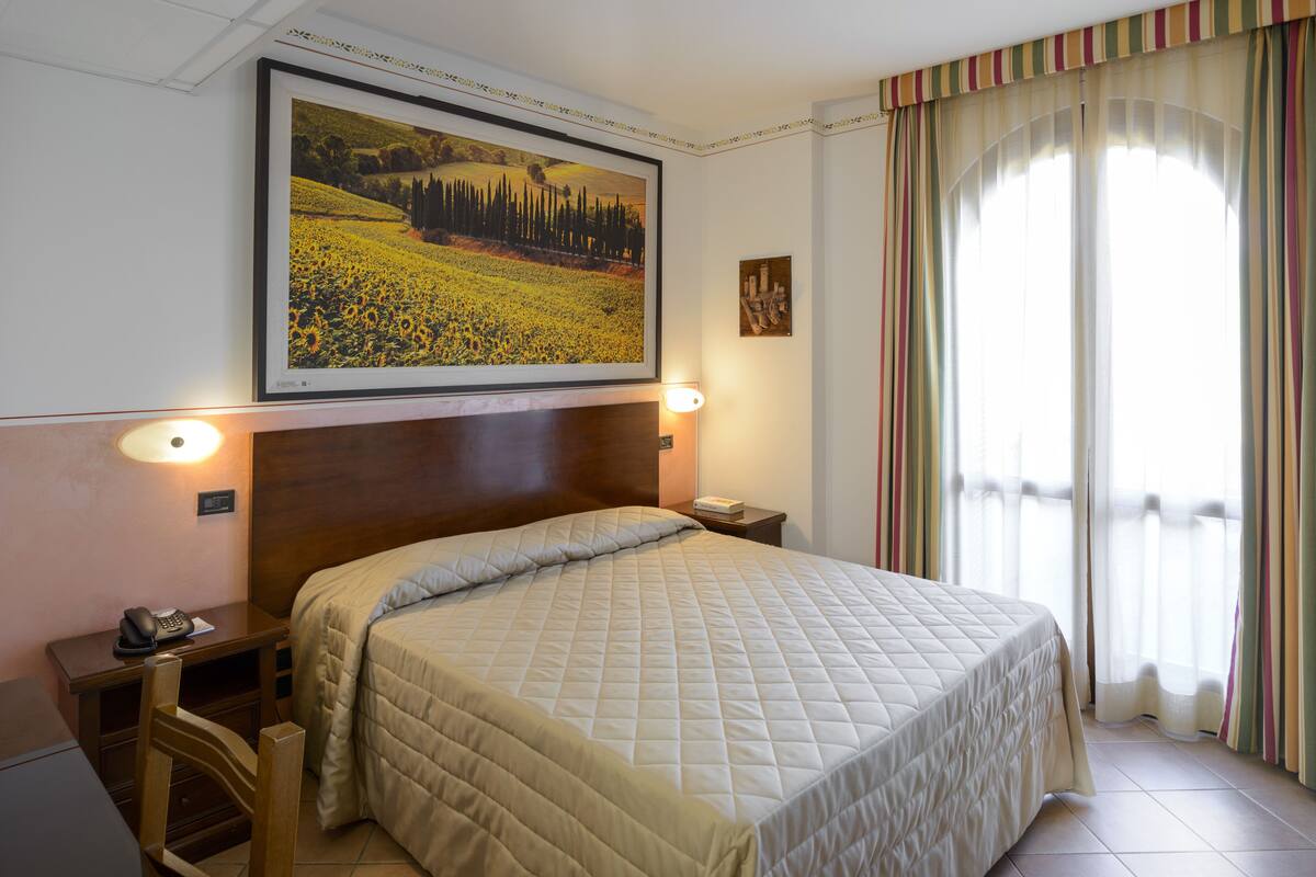 A hotel room with a large bed, a desk with a telephone, a chair, and a painting on the wall.