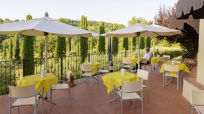 Un patio al aire libre con mesas amarillas y sombrillas blancas, rodeado por una barandilla metálica y vistas a un jardín ajardinado.