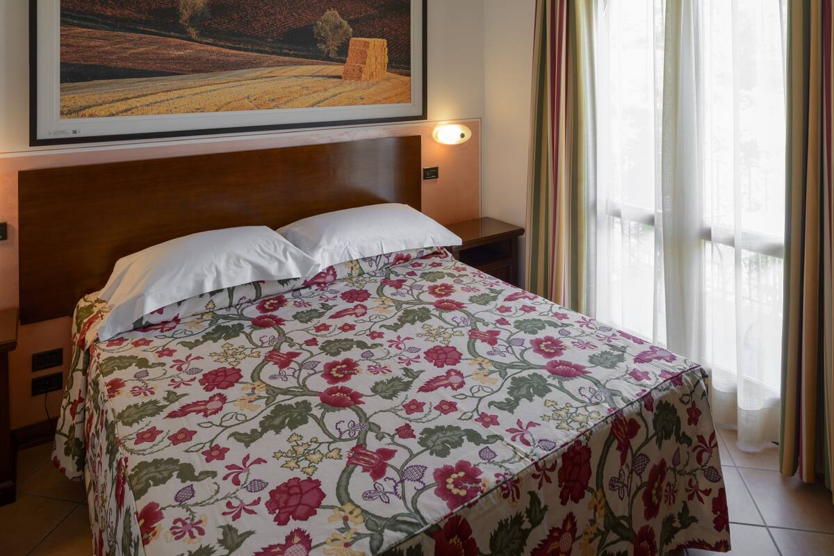 A hotel room with a bed, a painting on the wall, a nightstand, and a window with curtains.