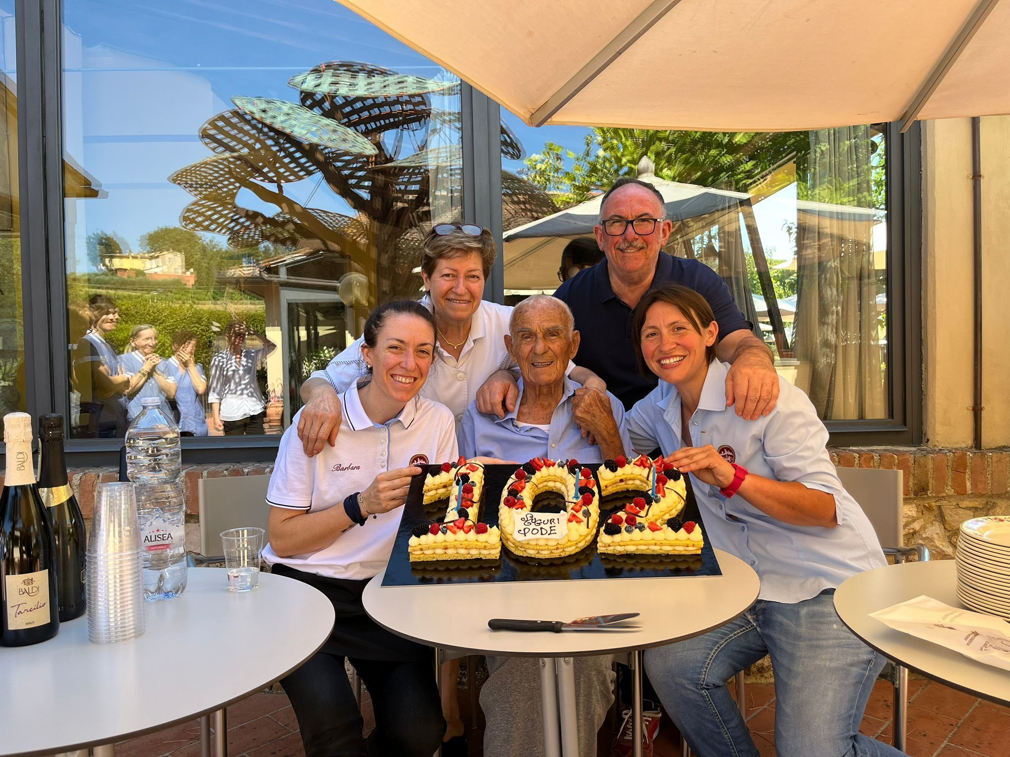 A group of people celebrating with a cake at an outdoor table with a view of a building and palm trees.