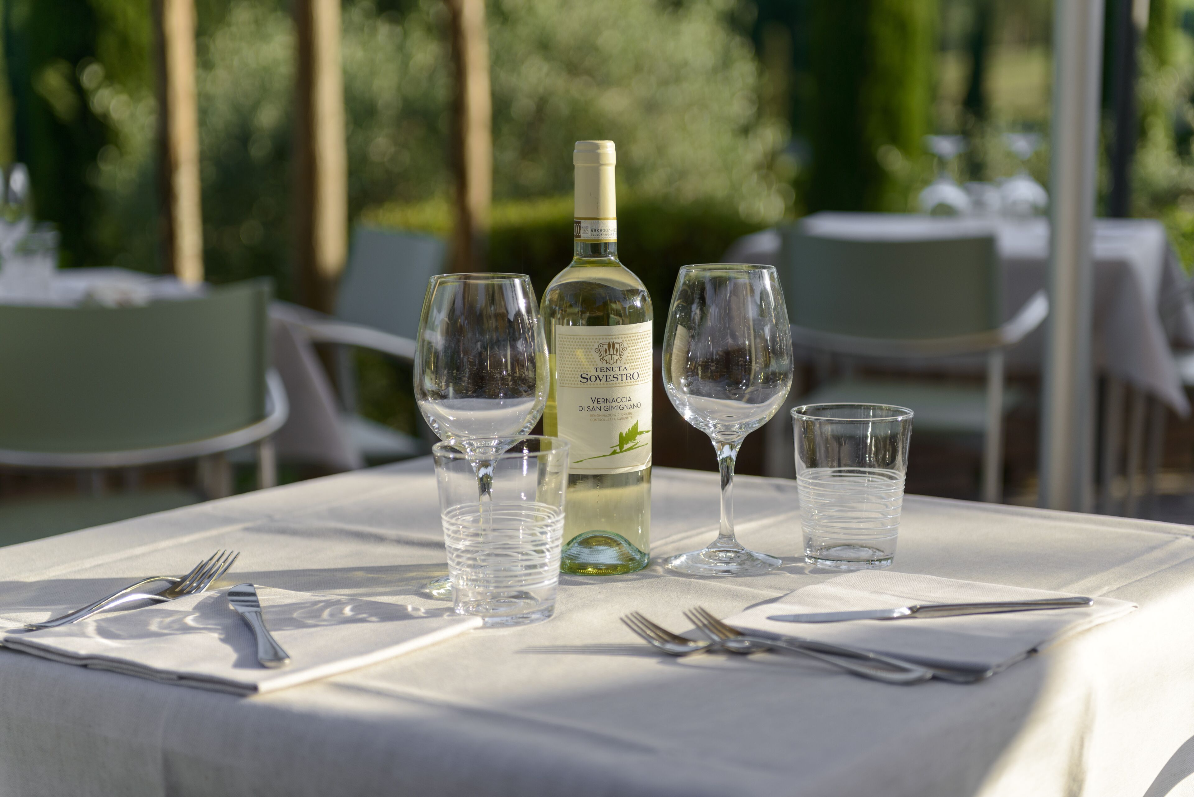 A table set for dining outdoors with a bottle of wine, glasses, and cutlery.
