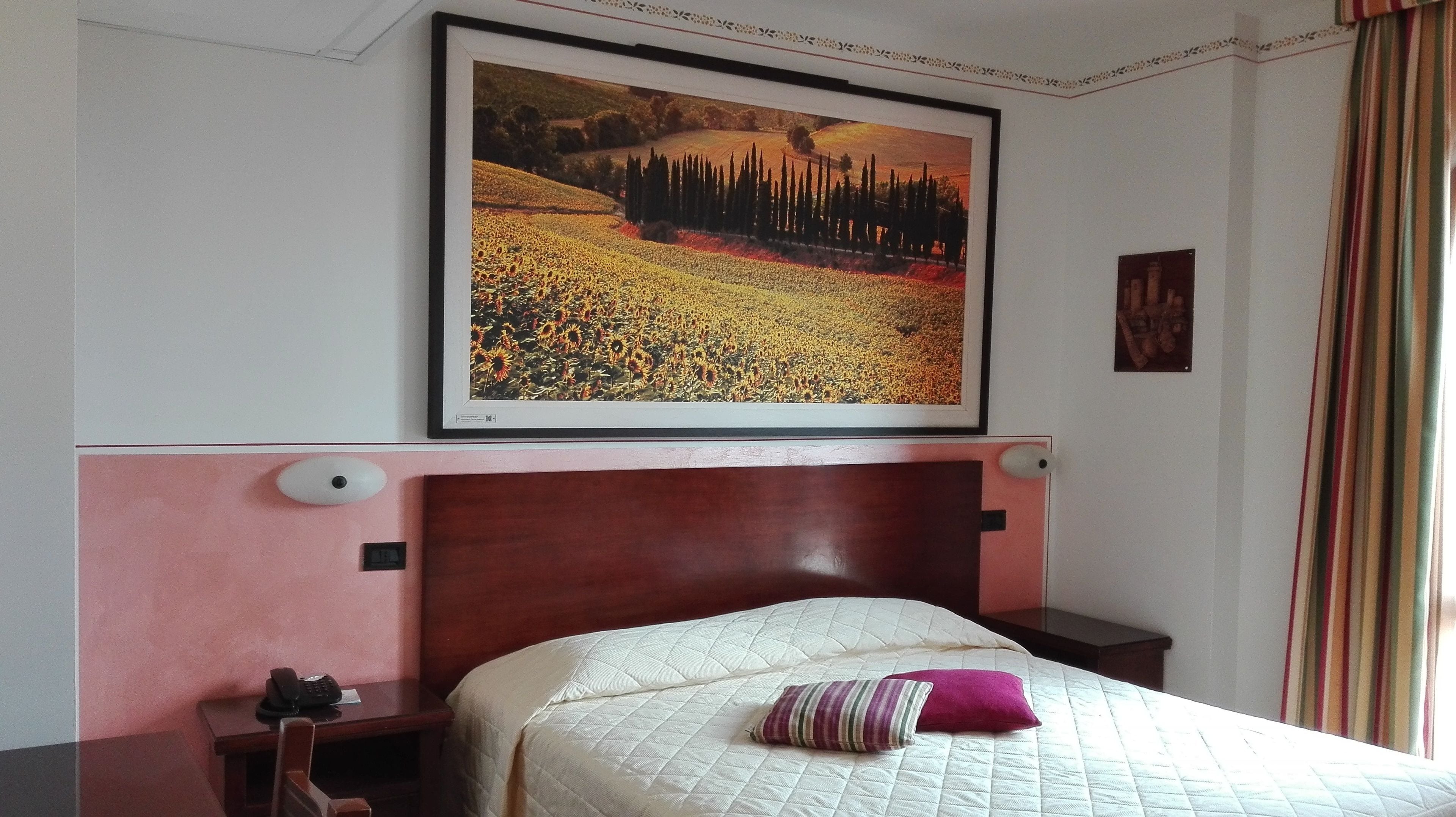 Quarto de hotel com cama, mesa de cabeceira, luminária, quadro decorativo de paisagem e janela com cortinas.