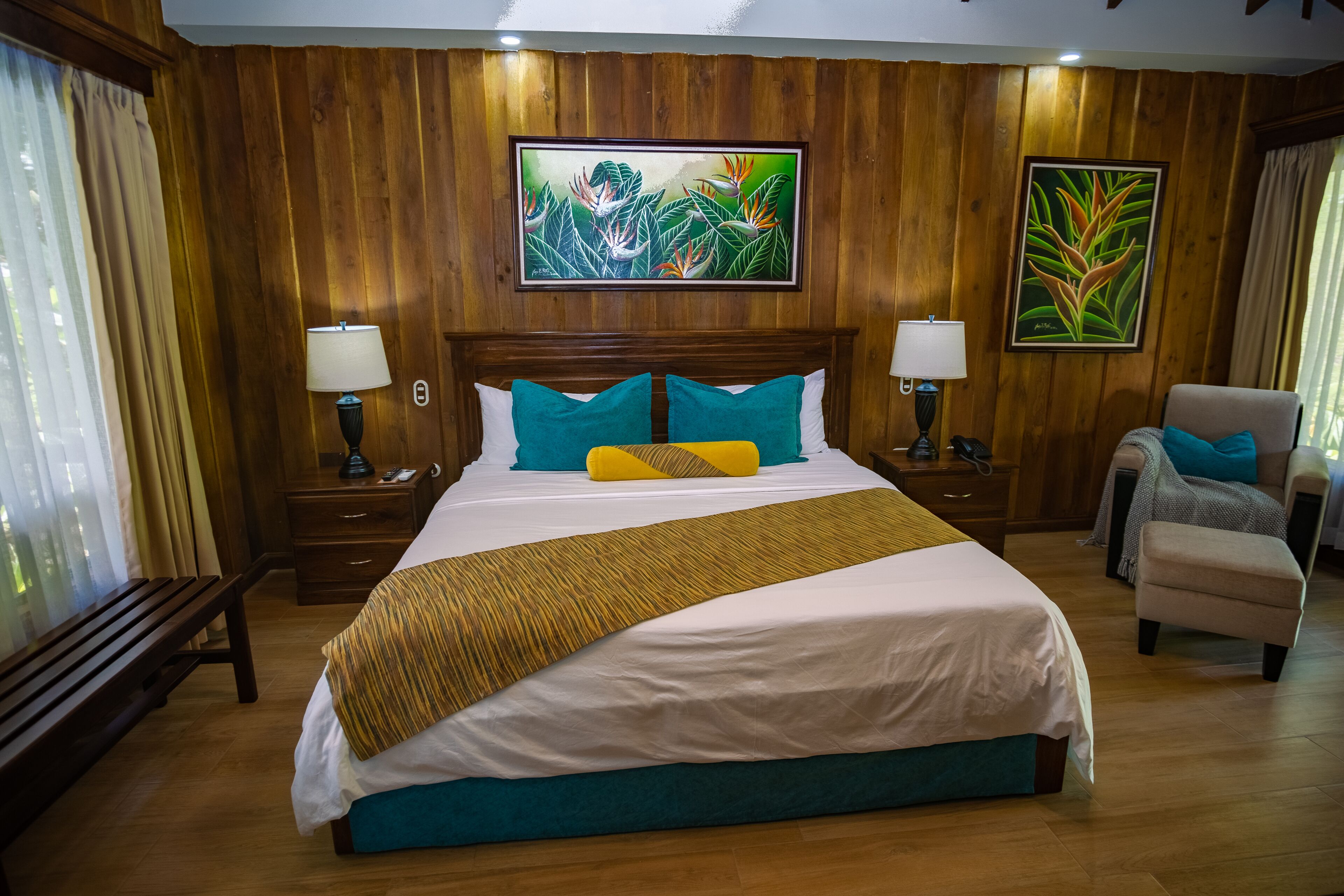 A hotel room with a large bed, wooden panel walls, two nightstands with lamps, a bench, a chair, and two paintings on the wall.