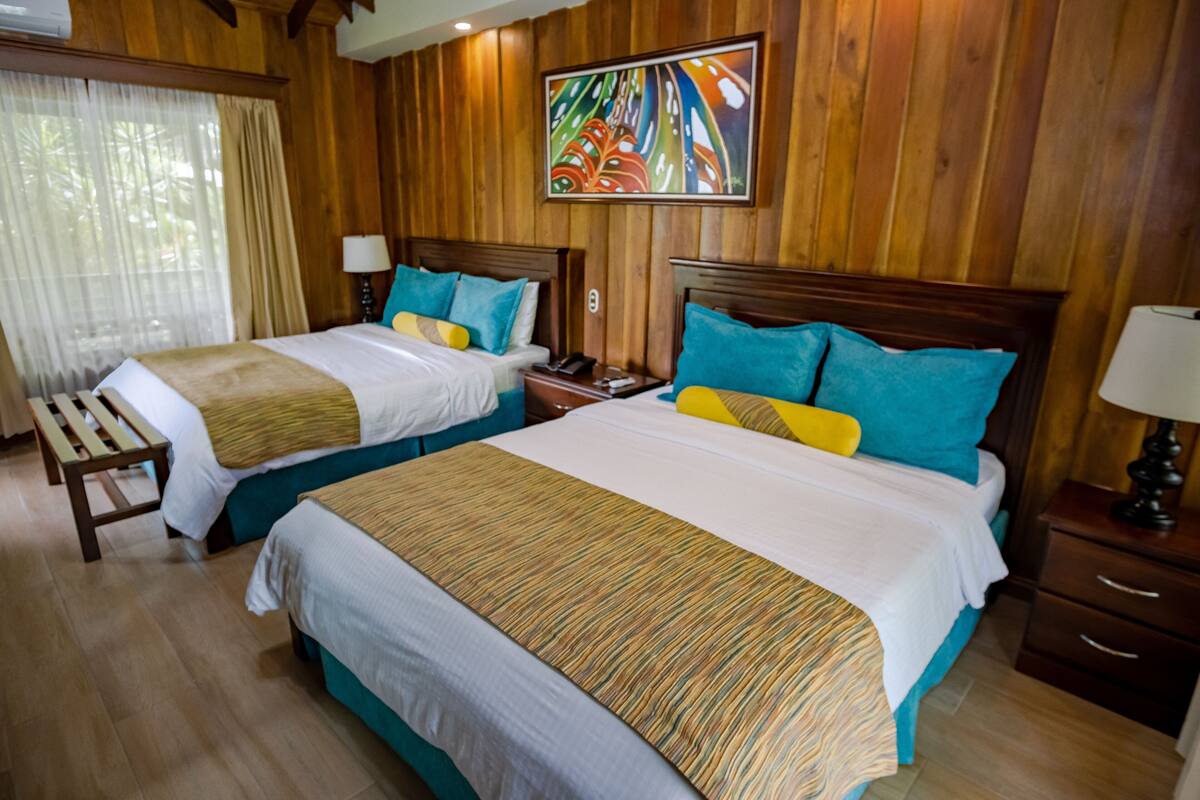 A hotel room with two beds, wooden walls, a painting, and a bench.