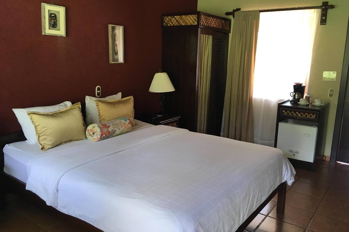 A hotel room with a bed, pillows, a nightstand with a lamp, a coffee maker, and a small refrigerator.