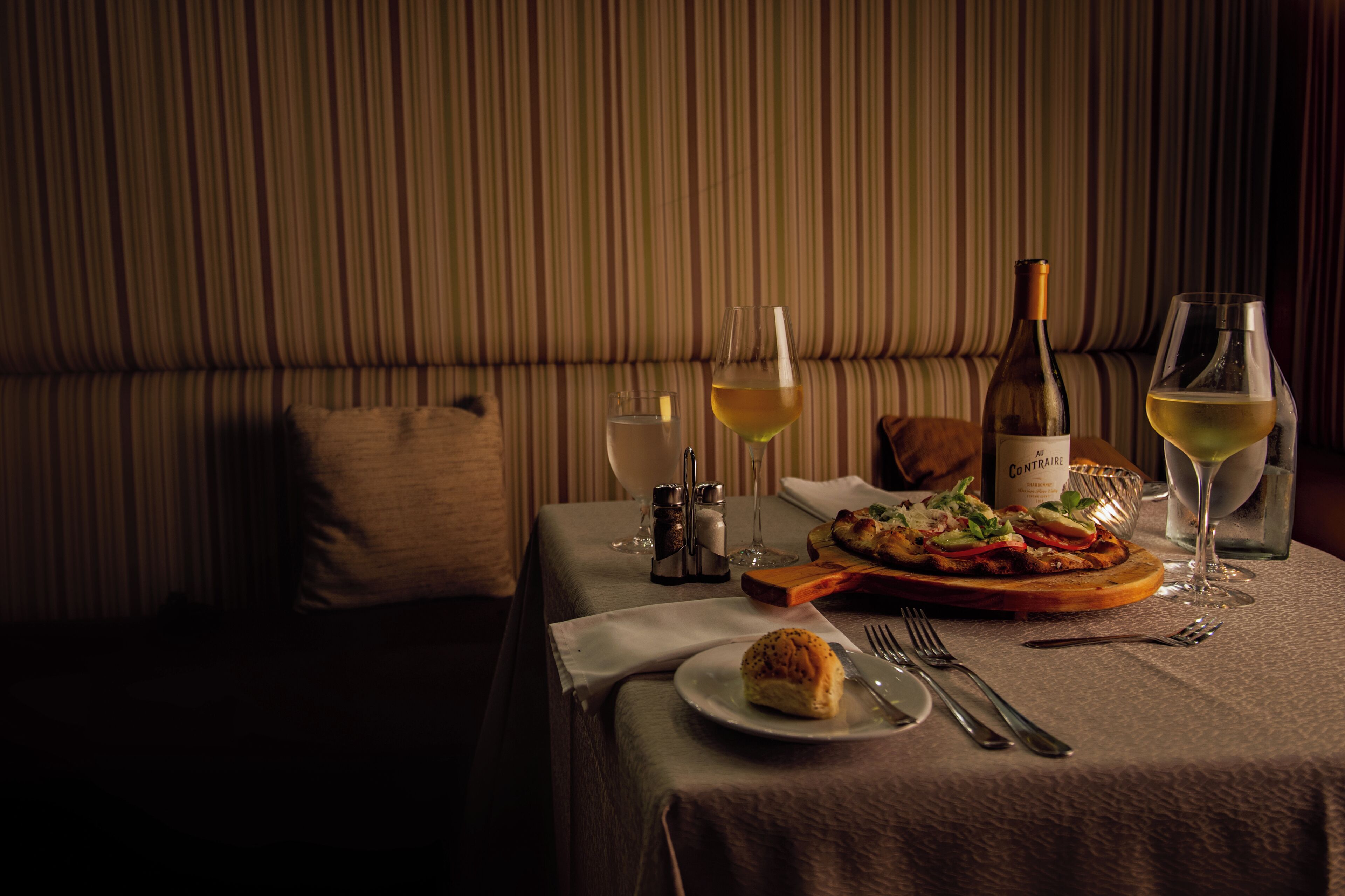 A dining table set with a plate of food, a bottle of wine, and two glasses of wine.