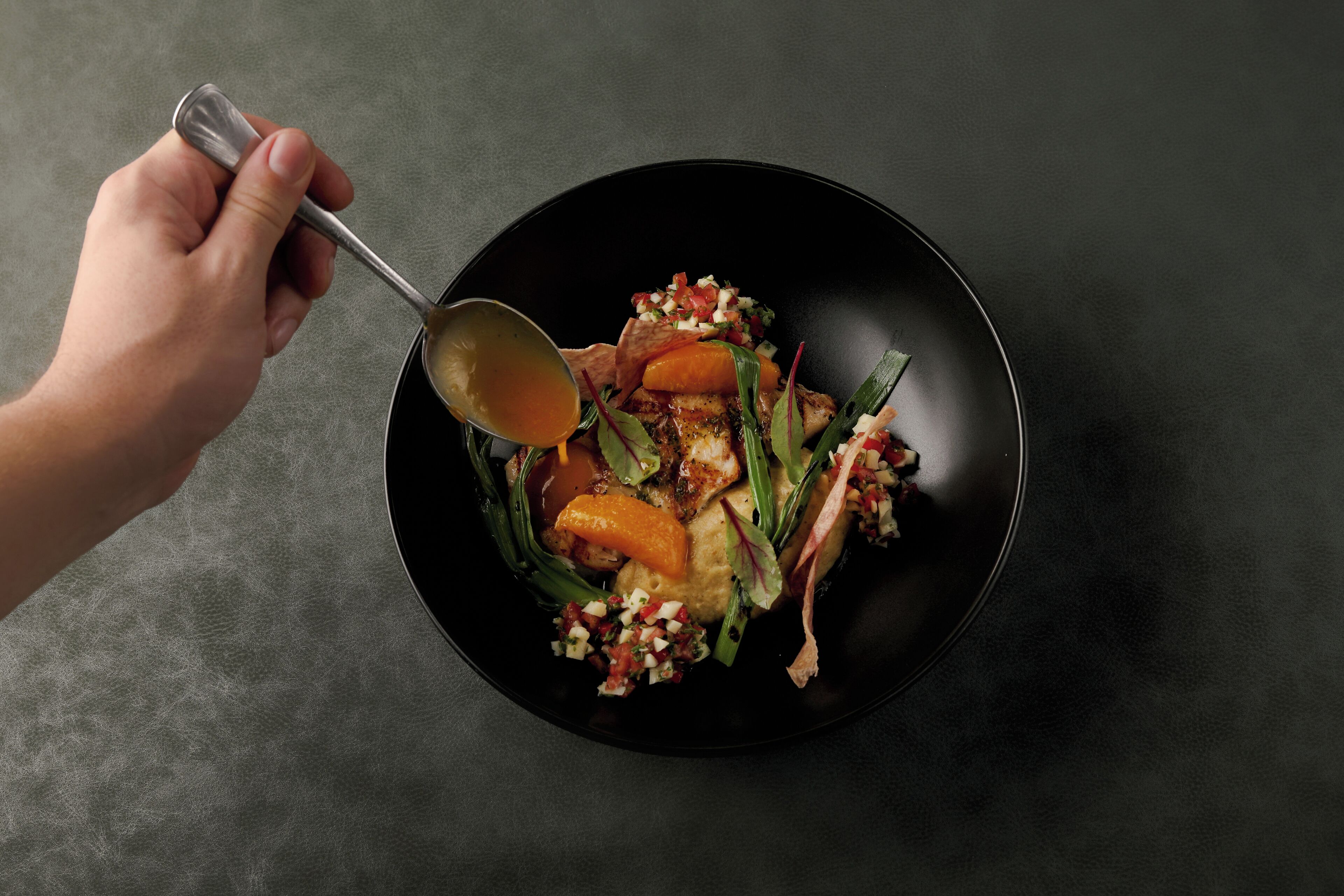 A hand holding a spoon over a black bowl containing a salad with various vegetables and a dressing.