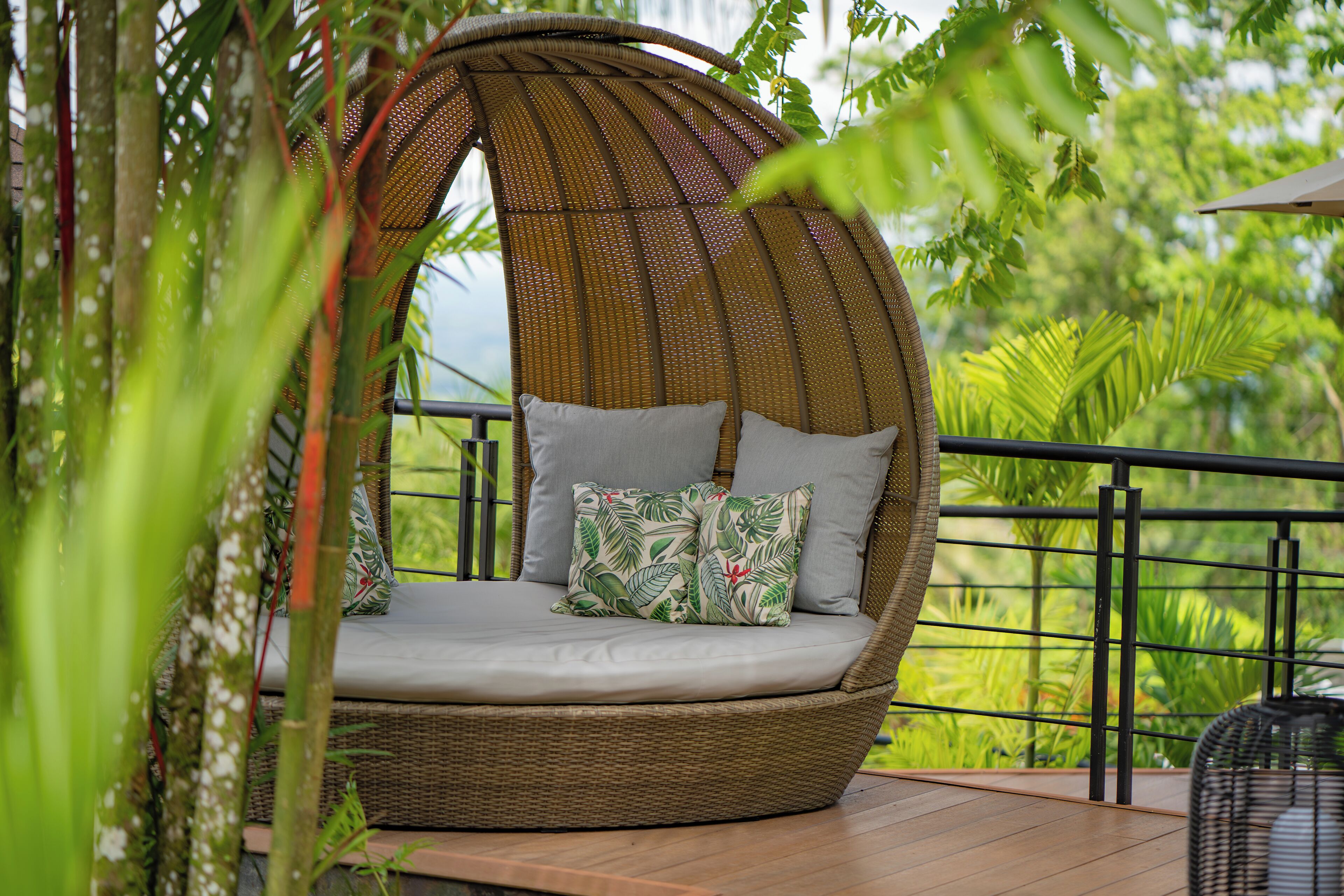 Un fauteuil suspendu en osier, doté de coussins, placé sur une terrasse en bois avec une rambarde en métal, et entouré d’une végétation luxuriante.