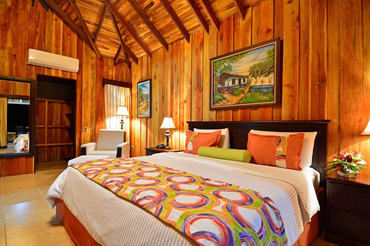 A wooden-paneled room with a bed, two chairs, a desk, and a painting on the wall.