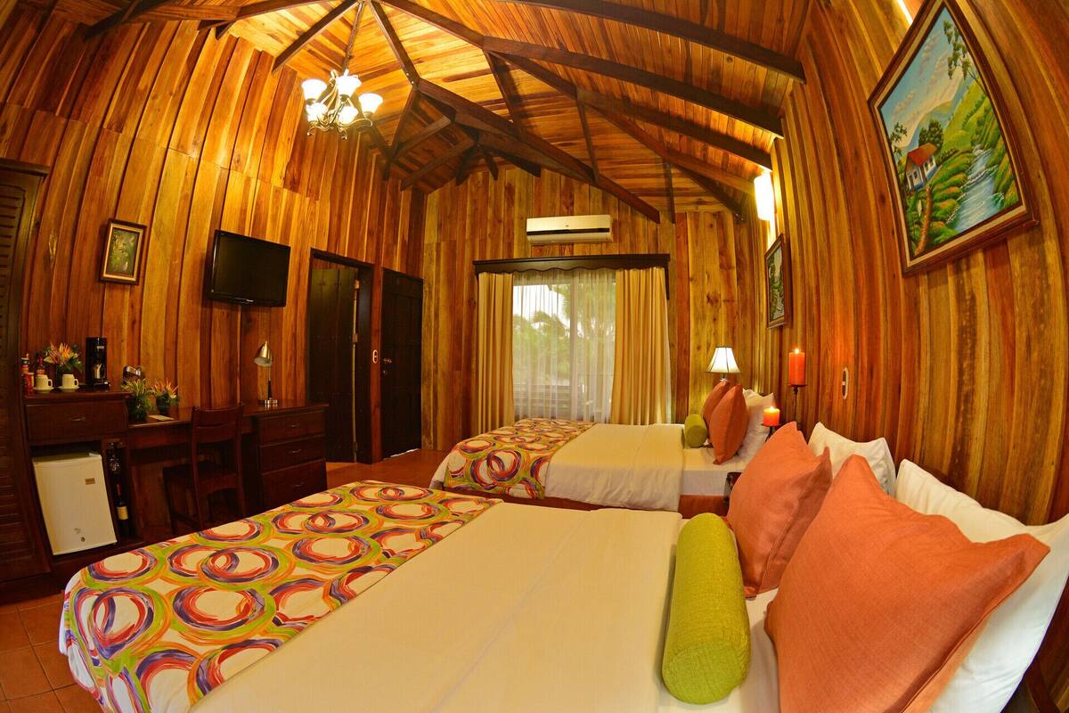 A room with wooden walls, a bed with colorful bedding, a desk with a chair, a television, and a painting on the wall.