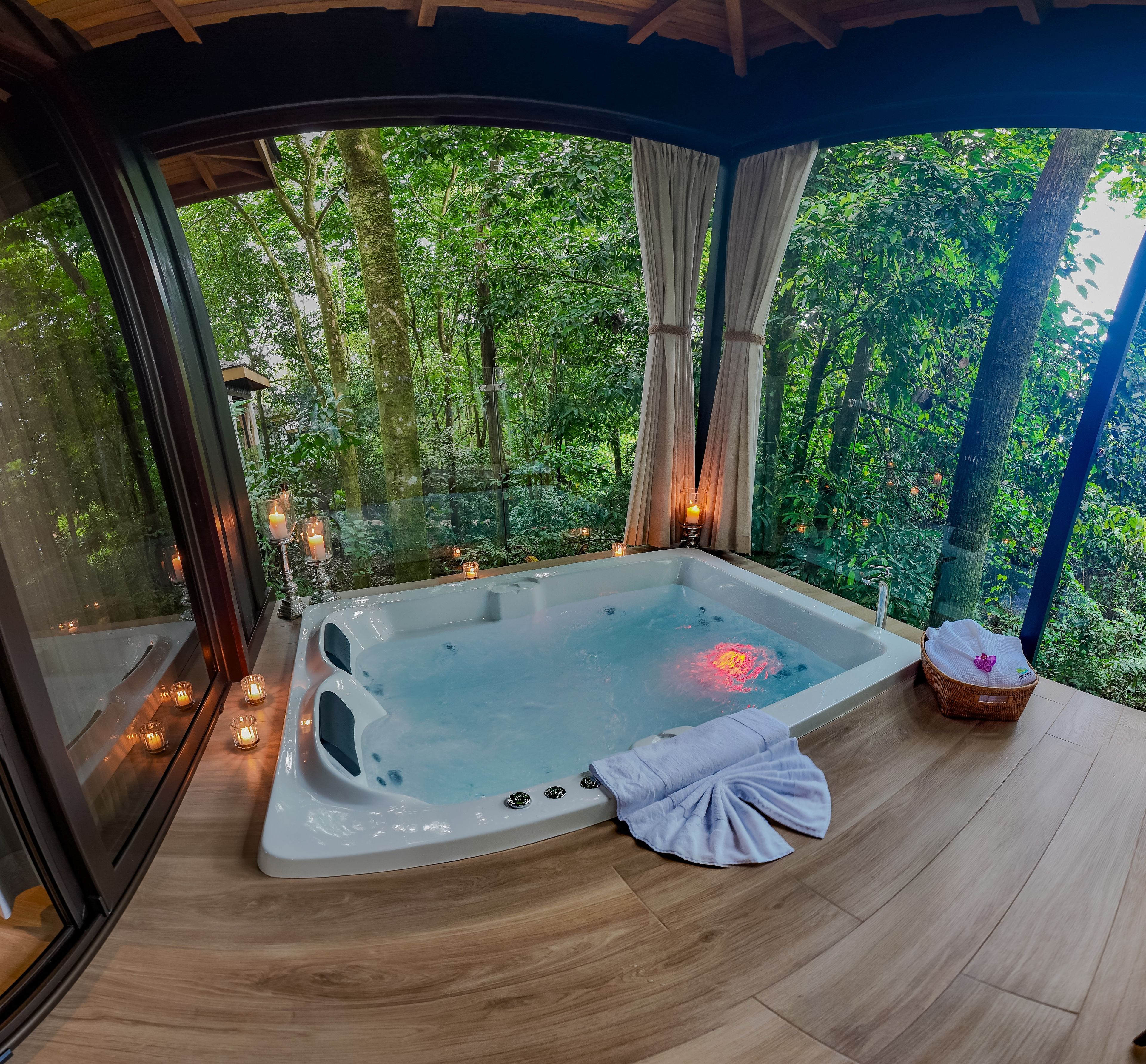 A hot tub with candles, a towel, and a basket on a wooden deck with a forest view.