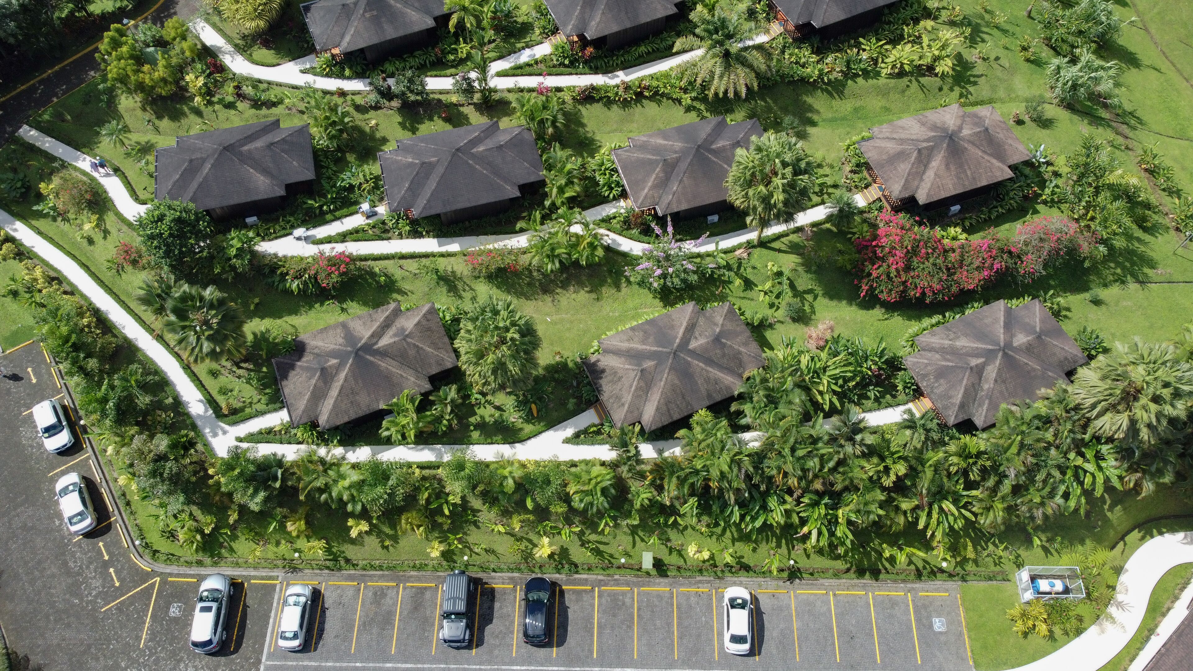 An aerial view of a resort with multiple bungalow-style buildings, arranged in a semi-circular pattern around a central landscaped area. There is a parking lot with several cars parked, and the resort is surrounded by lush greenery and palm trees.