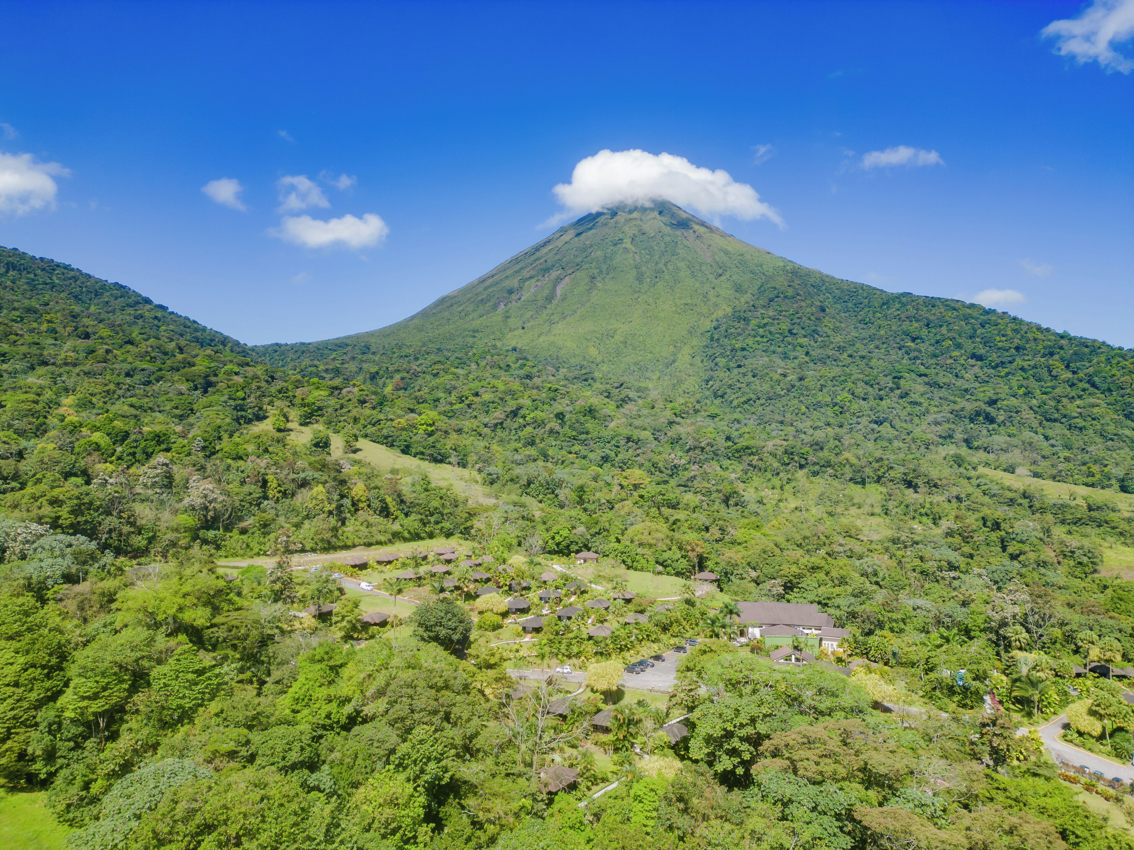 A lush green landscape with a prominent volcano in the background, surrounded by dense vegetation and a few scattered buildings.