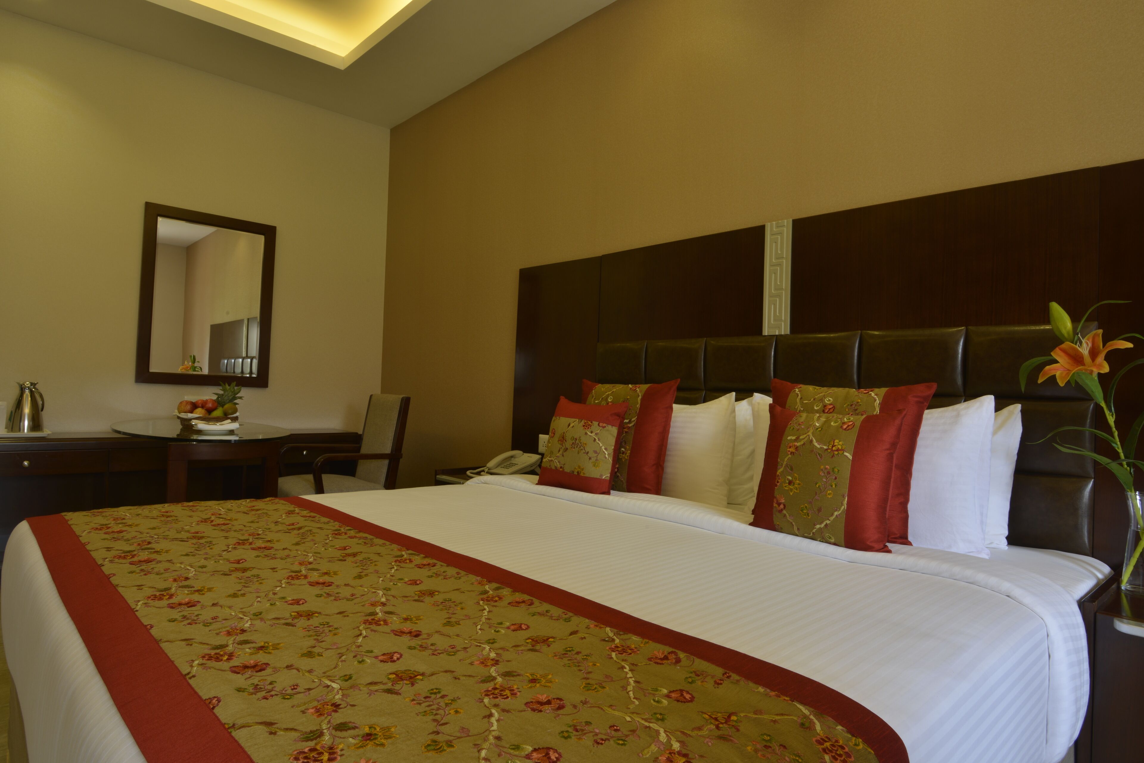 Ein Hotelzimmer mit einem großen Bett, einem Schreibtisch mit Telefon, einem Spiegel und einer Vase mit Blumen.