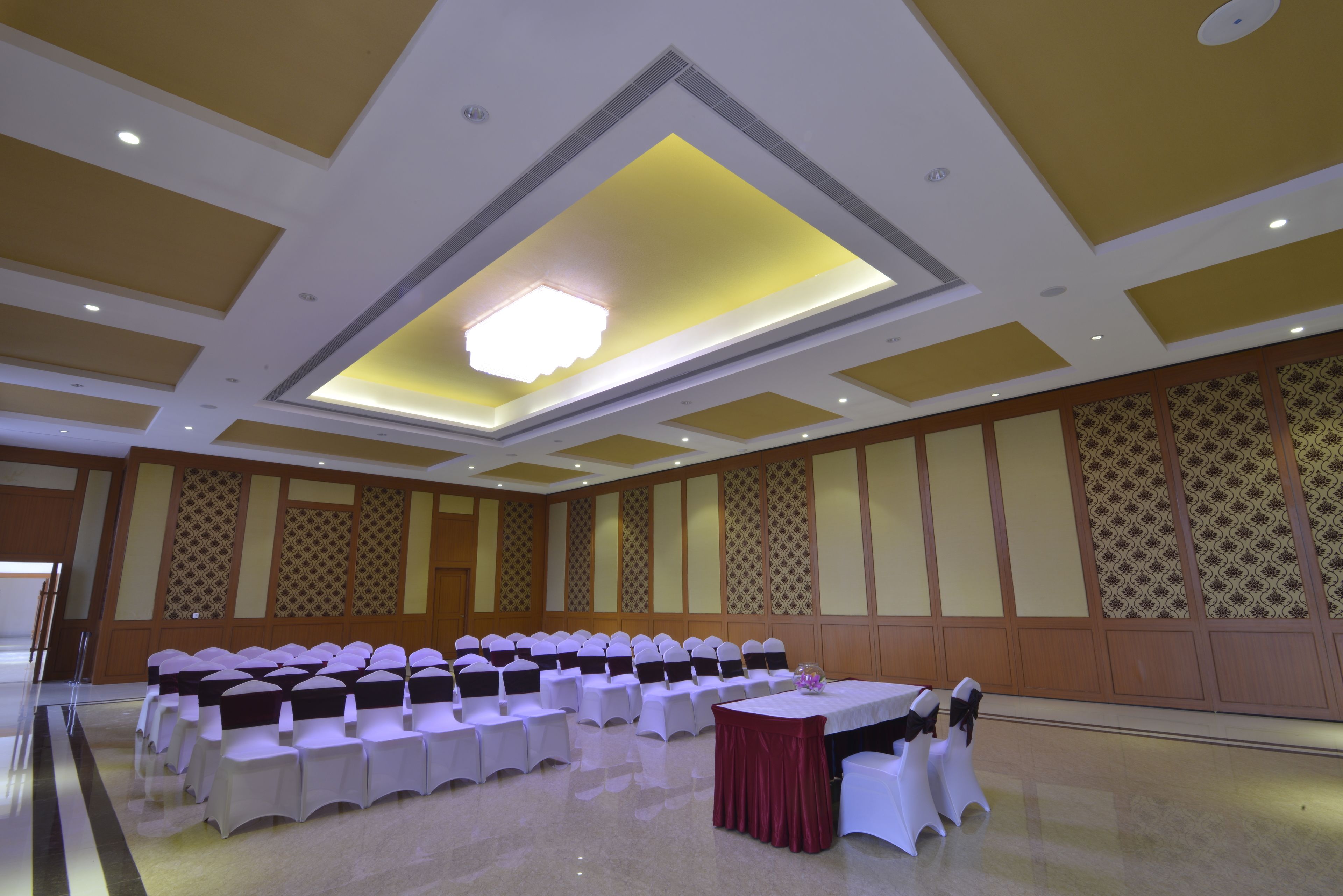 A conference room with rows of white tables and chairs, a central chandelier, and patterned wall panels.