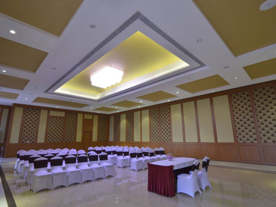 A conference room with rows of white tables and chairs, a central chandelier, and patterned wall panels.