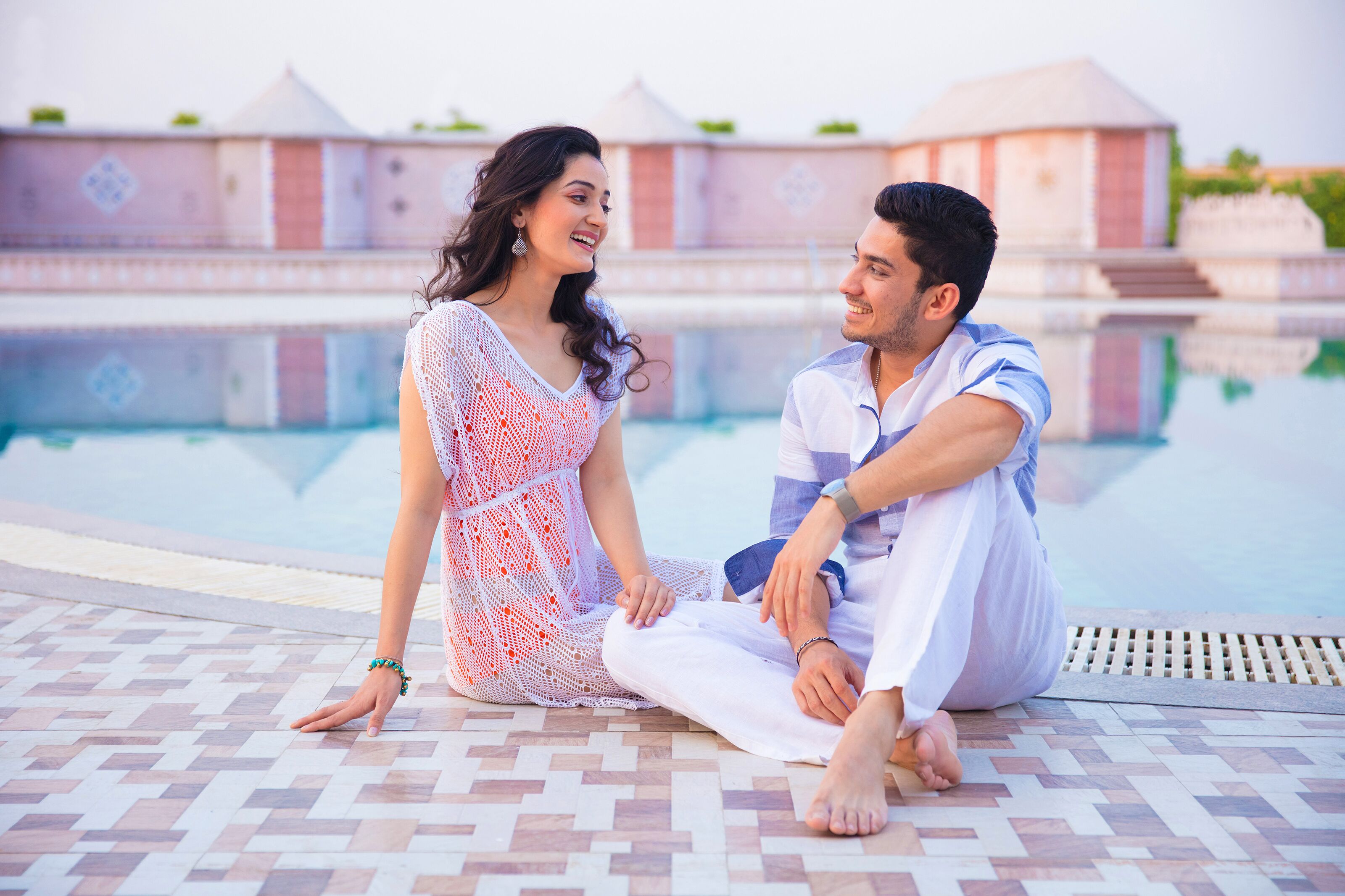A couple sitting by a pool with tiled flooring and a building in the background.