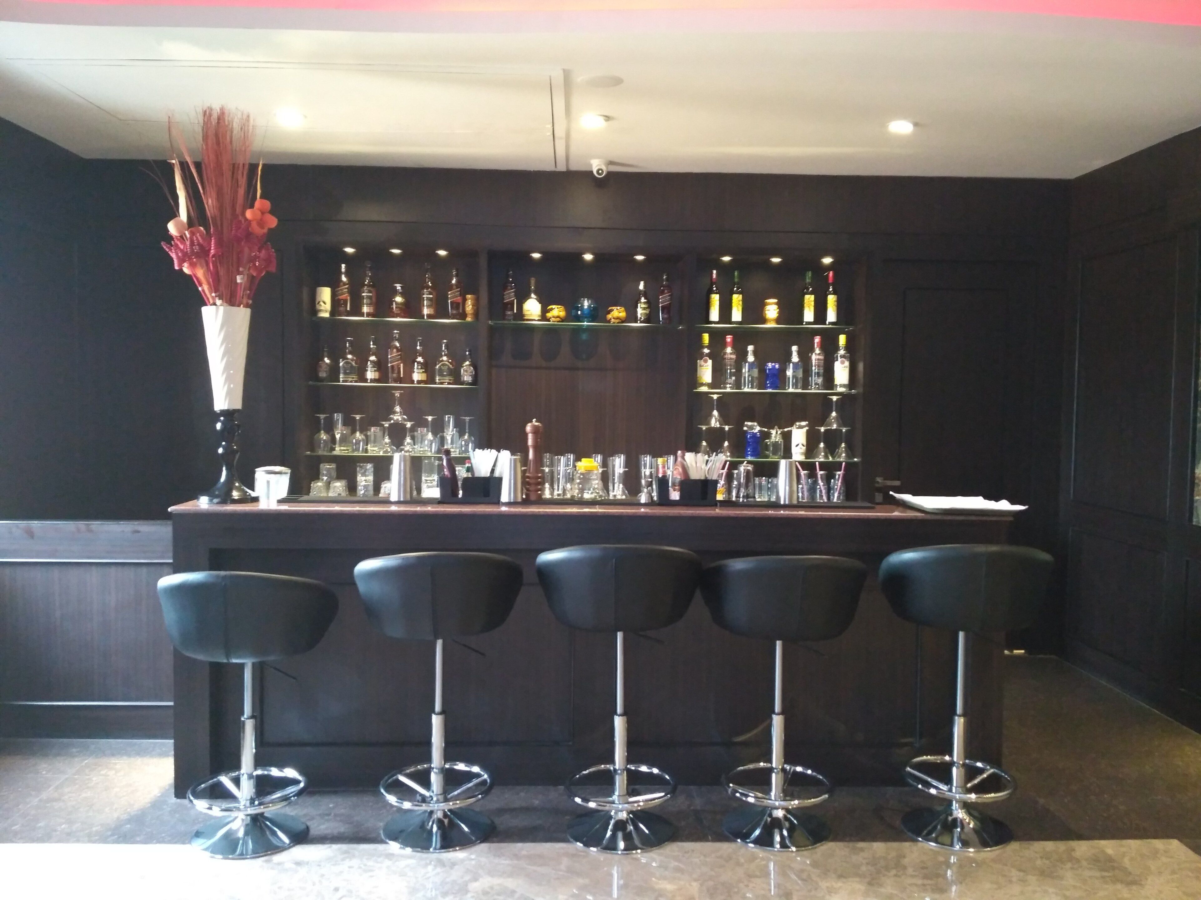 A bar area with a dark wood bar counter, multiple shelves of liquor bottles, and four black bar stools.