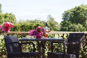 Uma mesa com duas cadeiras, uma garrafa e um copo de vinho numa varanda exterior com vista para vegetação.