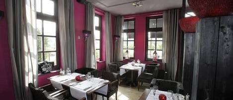 A restaurant with multiple tables set for dining, large windows with curtains, and a mix of wooden and upholstered chairs.