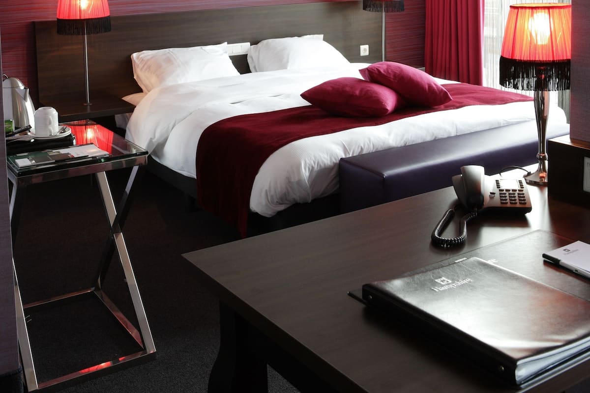 A hotel room with a bed, two bedside lamps, a desk with a phone, a lamp, and a framed picture on the wall.