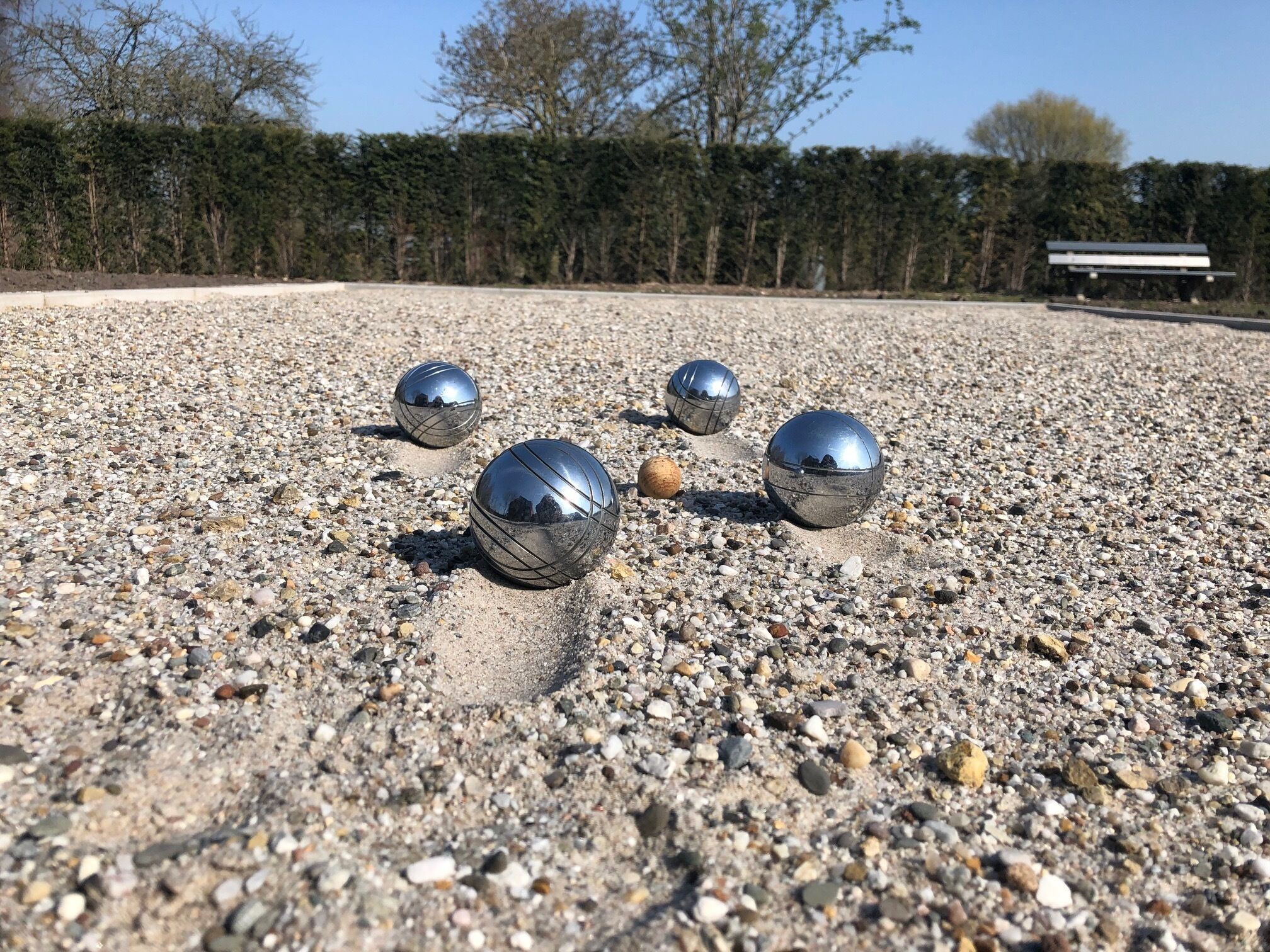Four metallic spheres on a gravel surface with a bench and trees in the background.