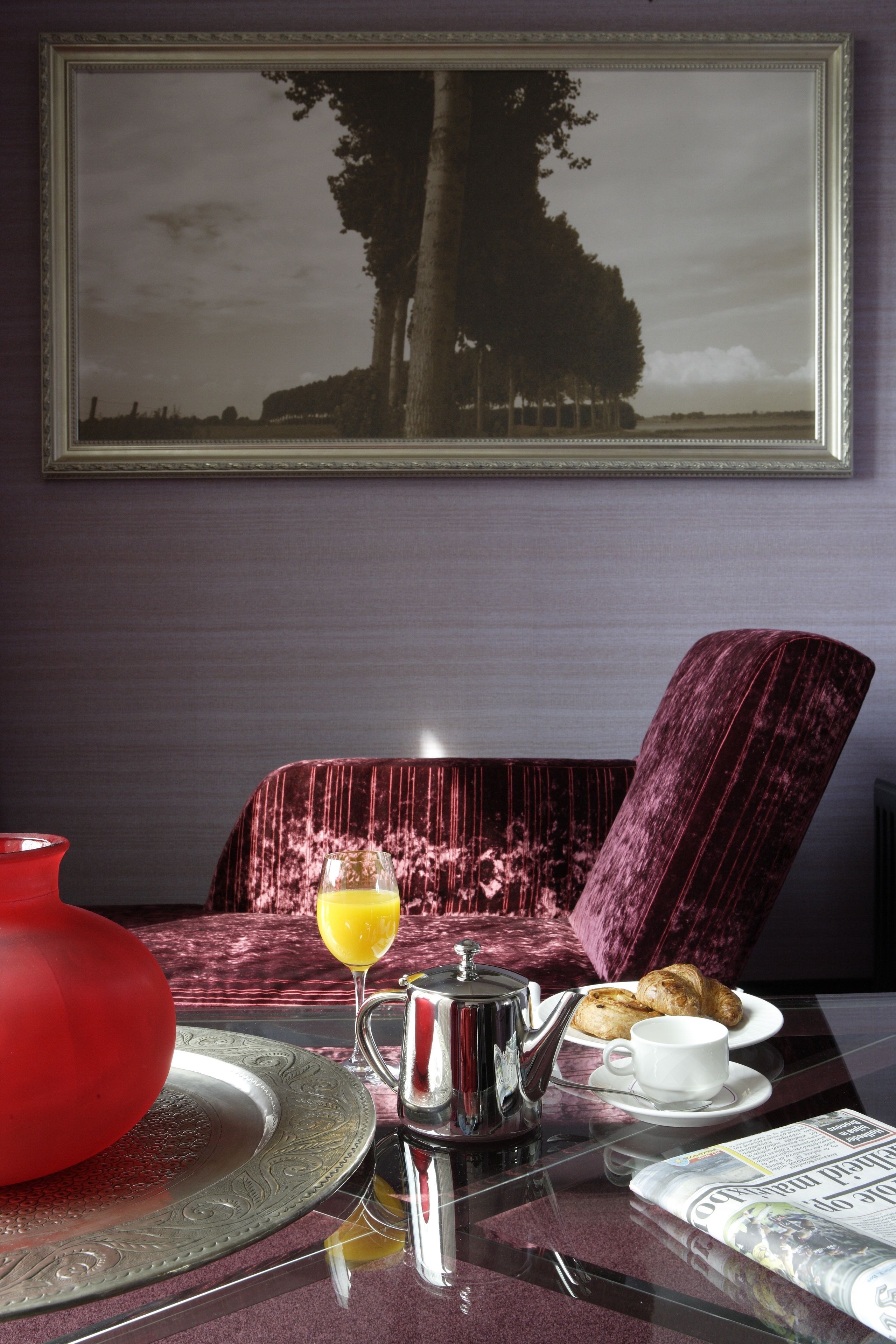 A dining table with a teapot, a red vase, a glass of orange juice, croissants, and a newspaper.