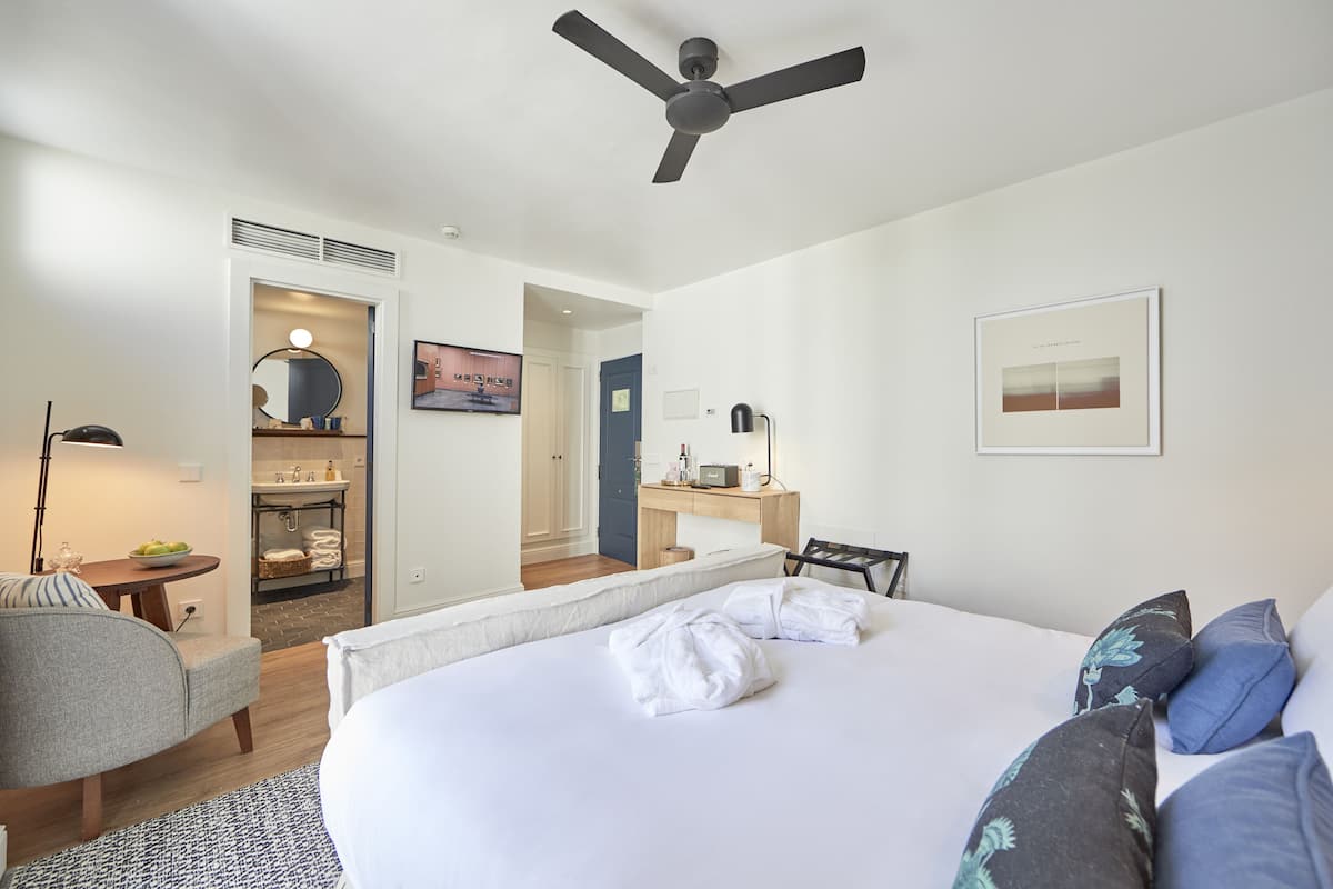 Une chambre d’hôtel avec un lit, un bureau, une chaise, un miroir, un ventilateur de plafond et une salle de bain visible à travers une porte ouverte.