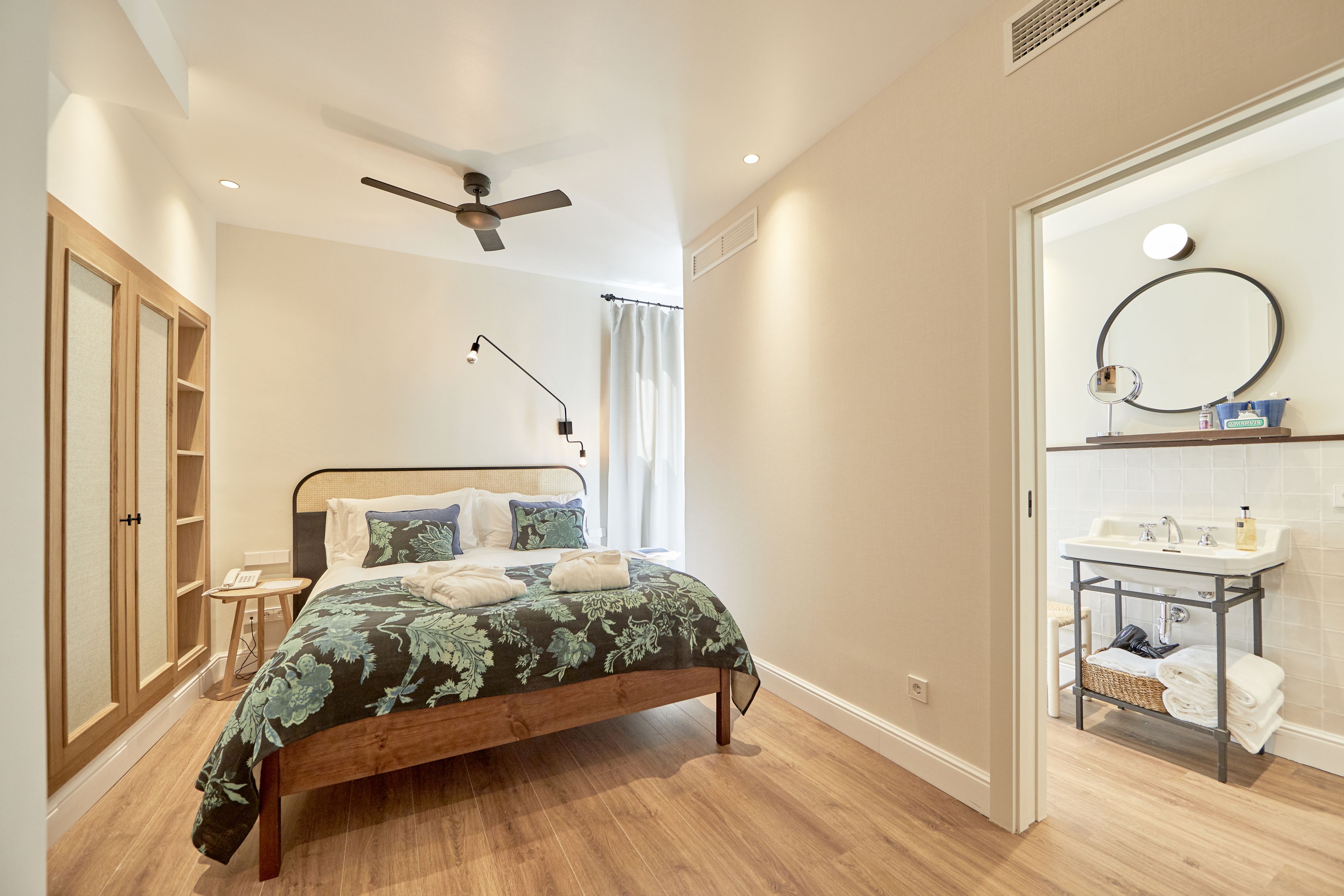 A bedroom with a wooden bed, a ceiling fan, a bedside table, and a bathroom with a sink and a mirror.