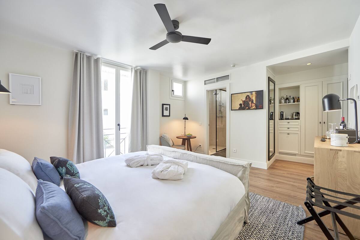 Une chambre d’hôtel avec un grand lit, un ventilateur de plafond, un bureau avec une lampe, une salle de bain avec douche et un coin repas avec une table et des chaises.