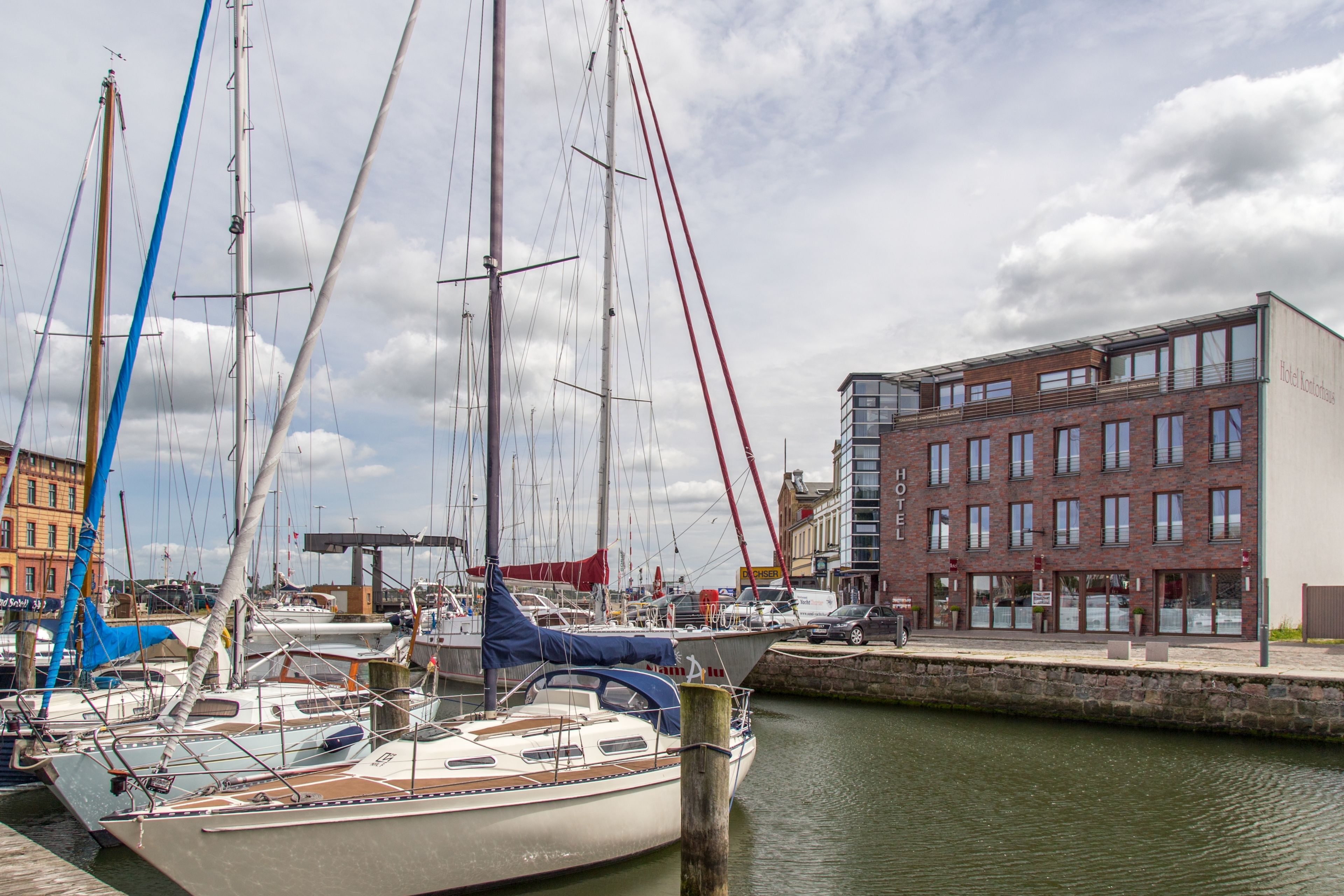 A marina with several sailboats docked, a hotel building with multiple windows, and a stone wall separating the water from the dock.
