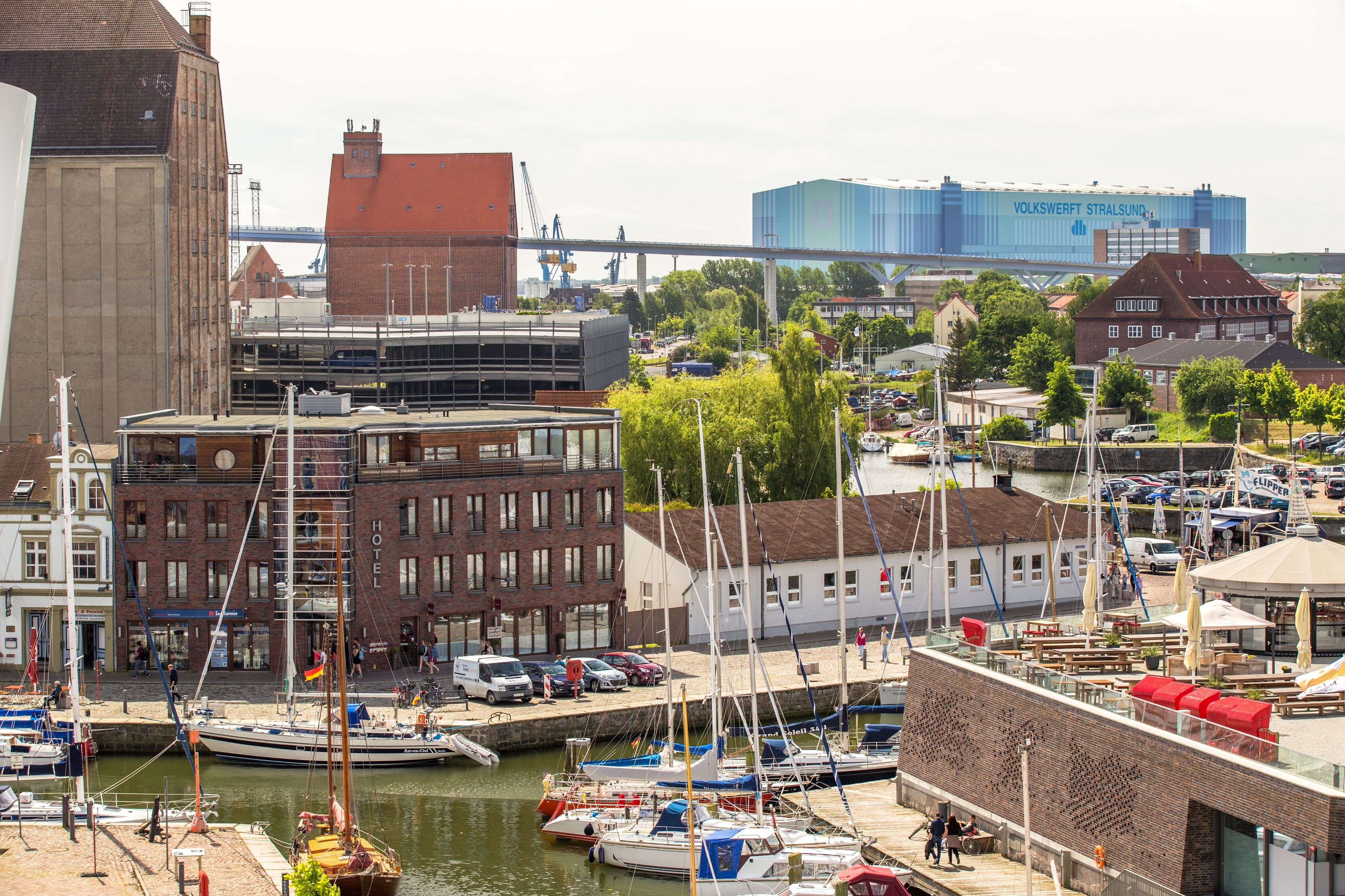 Un port de plaisance avec des bateaux amarrés, un hôtel et, au loin, un grand bâtiment industriel.