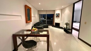 A modern living room with a glass dining table, black chairs, a black sofa, a fireplace, and a large window overlooking a cityscape at night. - 104 Art Suites (Bogotá)