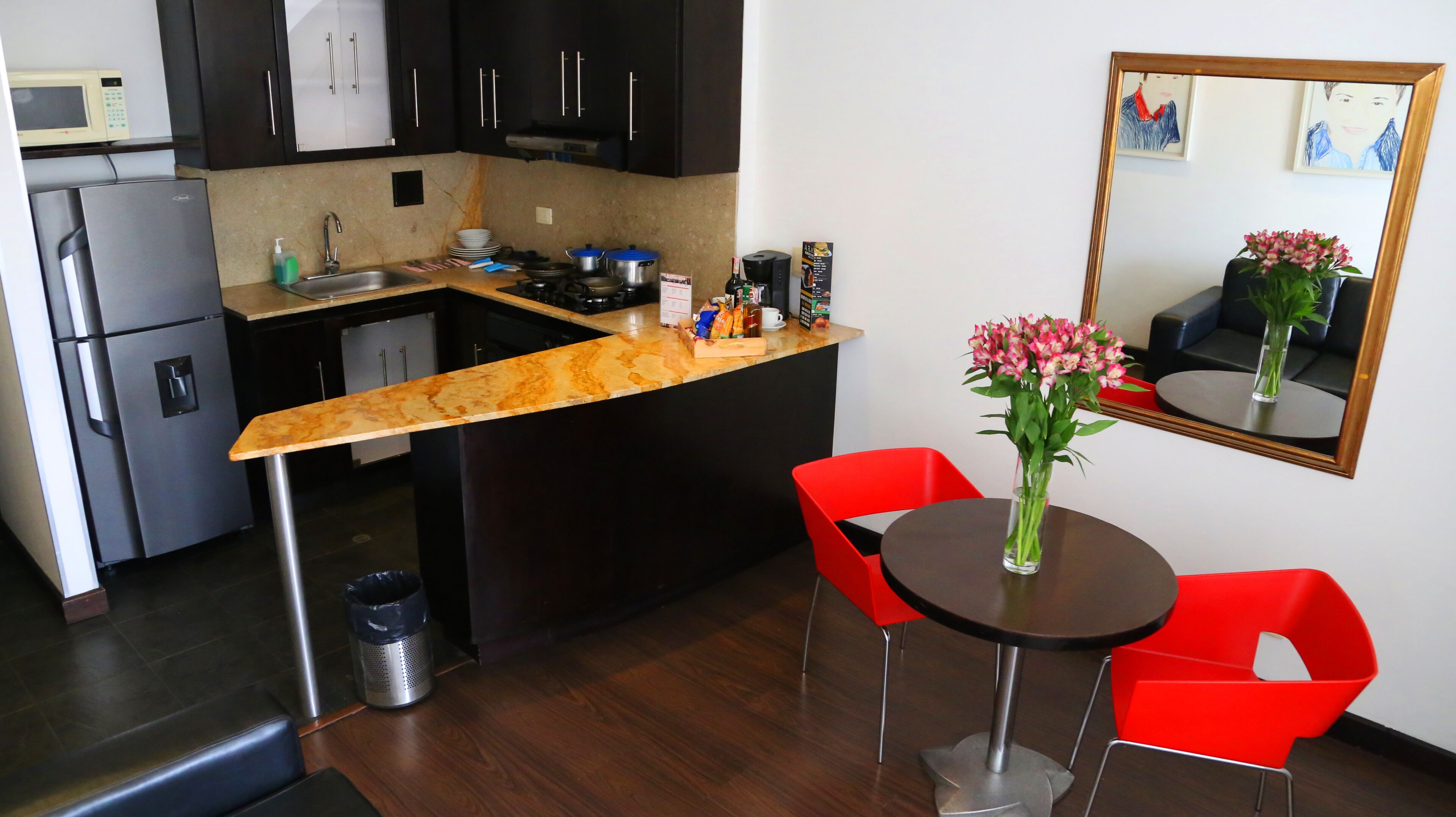 Een functionele keuken met een magnetron, koelkast en spoelbak. Er is een klein eethoekje met een tafel en twee rode stoelen. Op de tafel staat een vaas met bloemen en aan de muur hangt een spiegel.